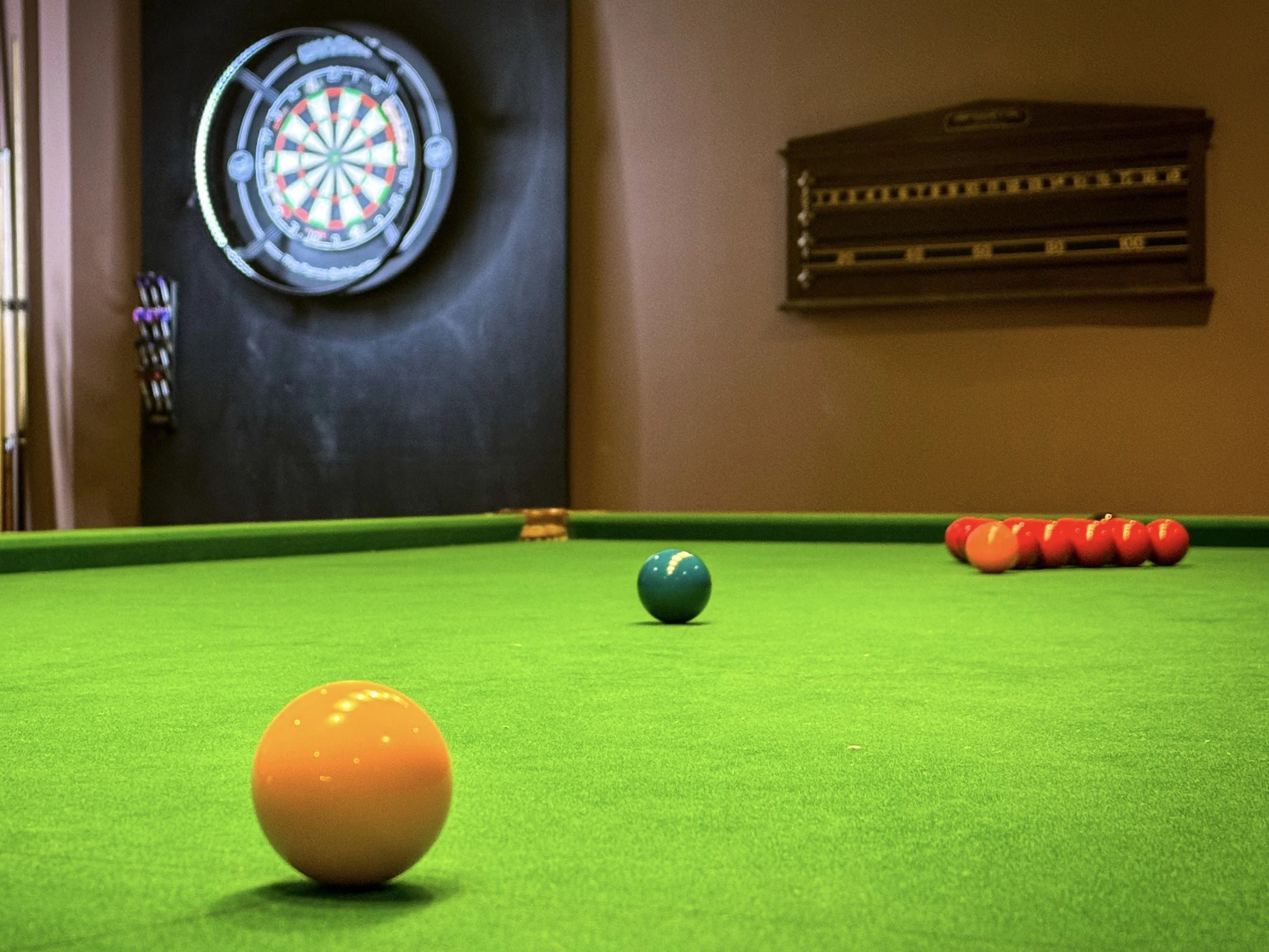 Colorful billiard balls on a green pool table with a dartboard on the wall in the background.