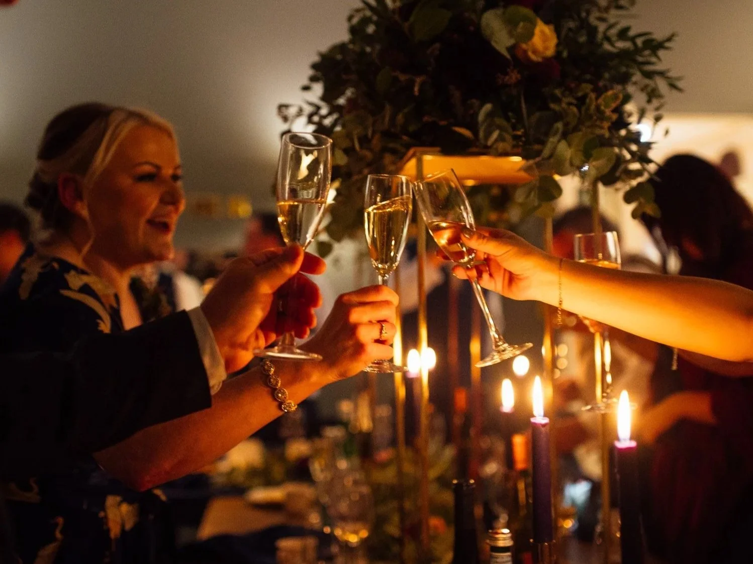 People clinking champagne glasses at a celebration or party in a warmly lit setting decorated with candles and flowers.