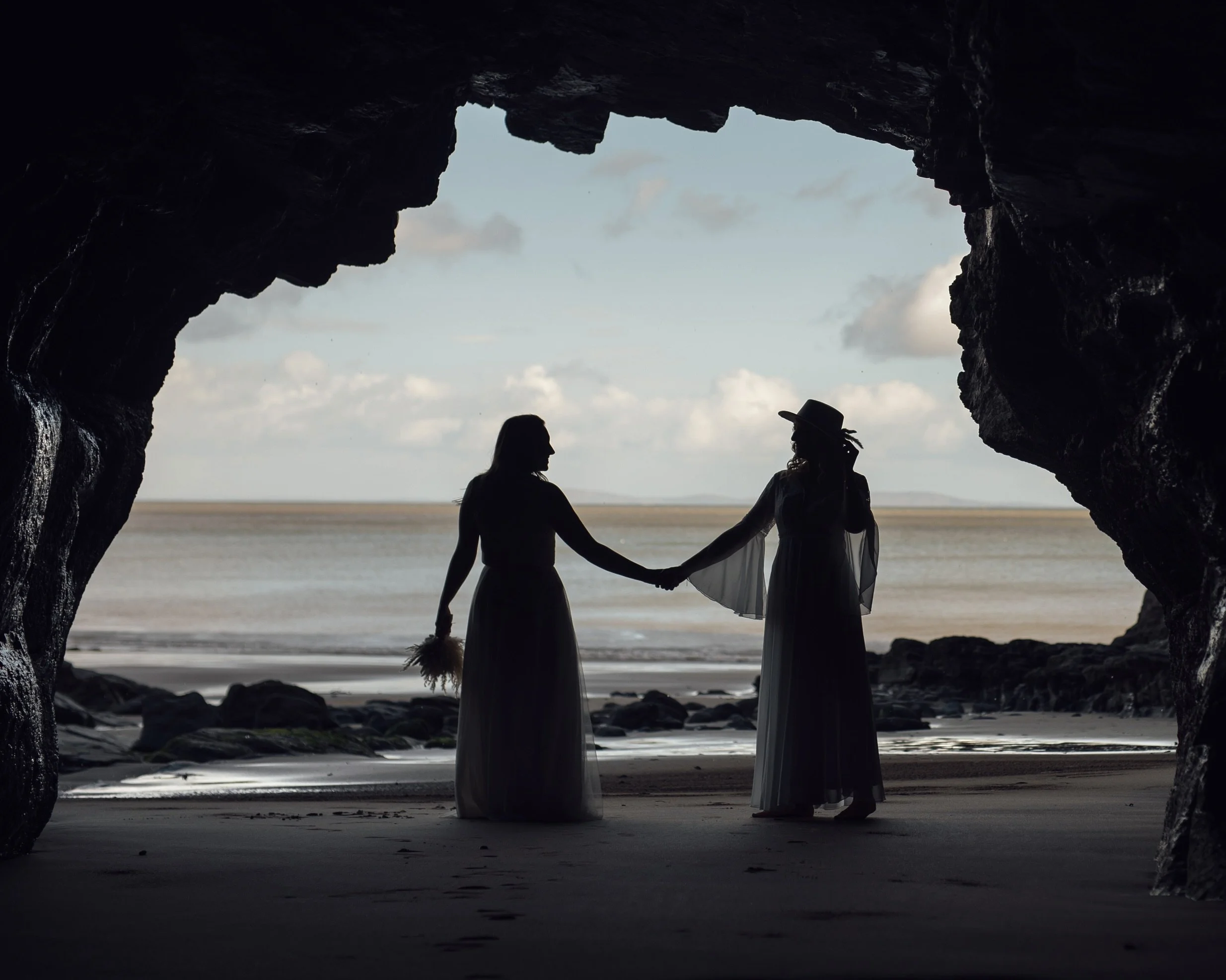 Two women holding hands inside a rocky seaside cave during sunset, with the ocean and sky visible in the background.