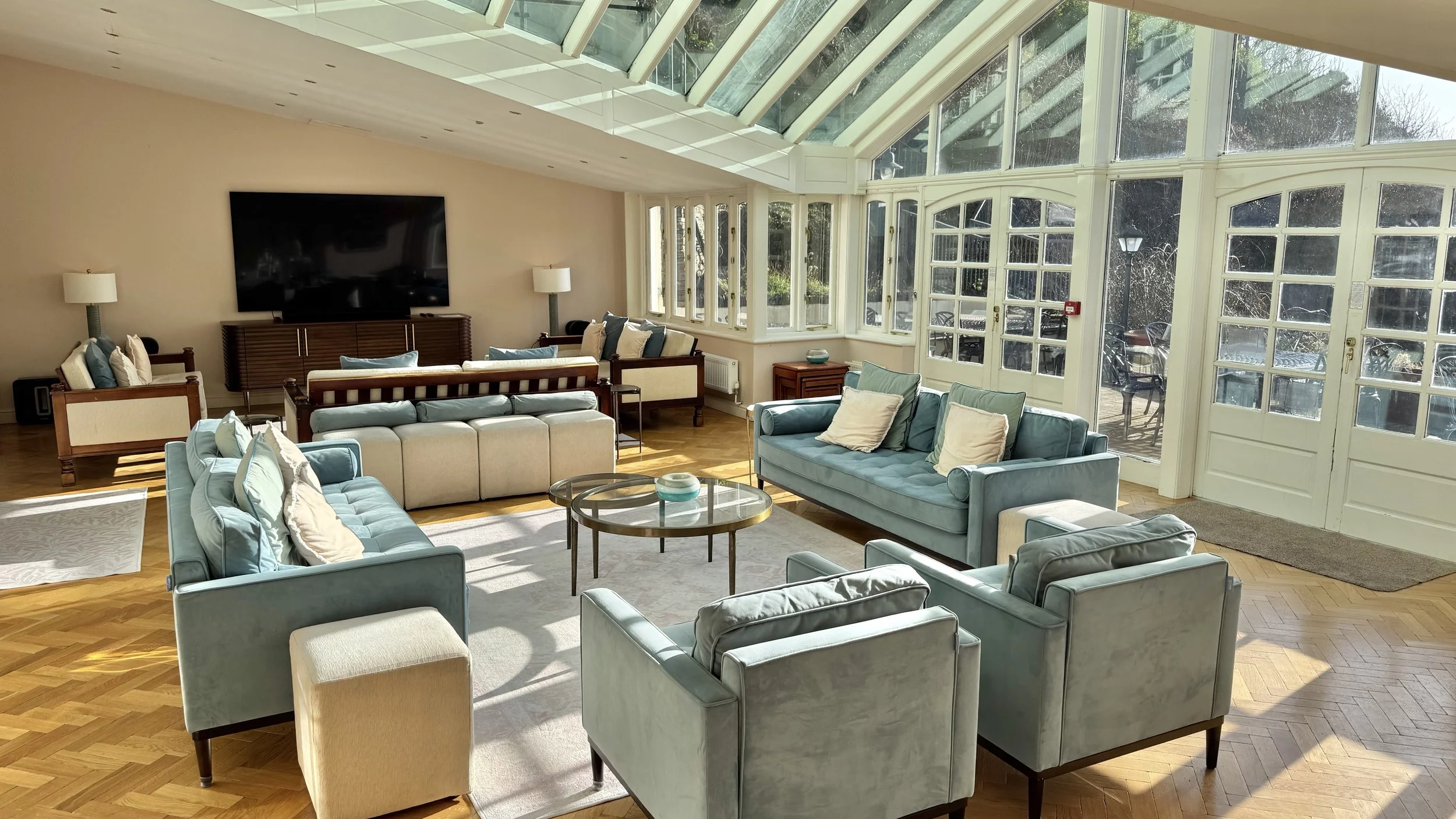 A spacious living room with large windows and a glass ceiling, furnished with blue and beige sofas, a flat-screen TV, and a wooden side table, decorated in a modern style with hardwood floors and natural light.