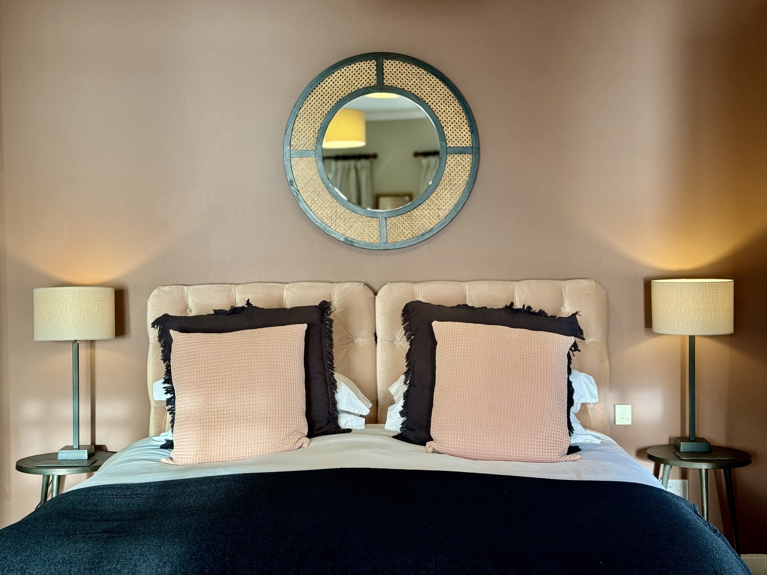 A bedroom with a quilted beige headboard, decorated with four pink and black pillows and white bedding. Two matching beige table lamps with green bases sit on black bedside tables. A round mirror with wicker detailing is mounted above the headboard, reflecting a yellow lamp and curtains.