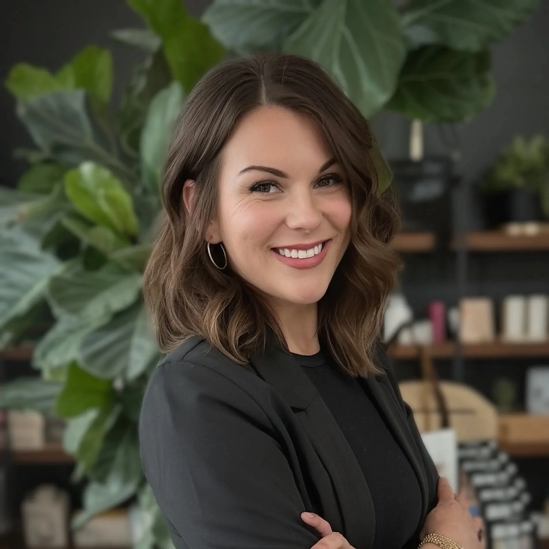 A woman with shoulder-length wavy brown hair, wearing a black blazer and hoop earrings, smiling with arms crossed in front of large green plants and shelves with books and decor.