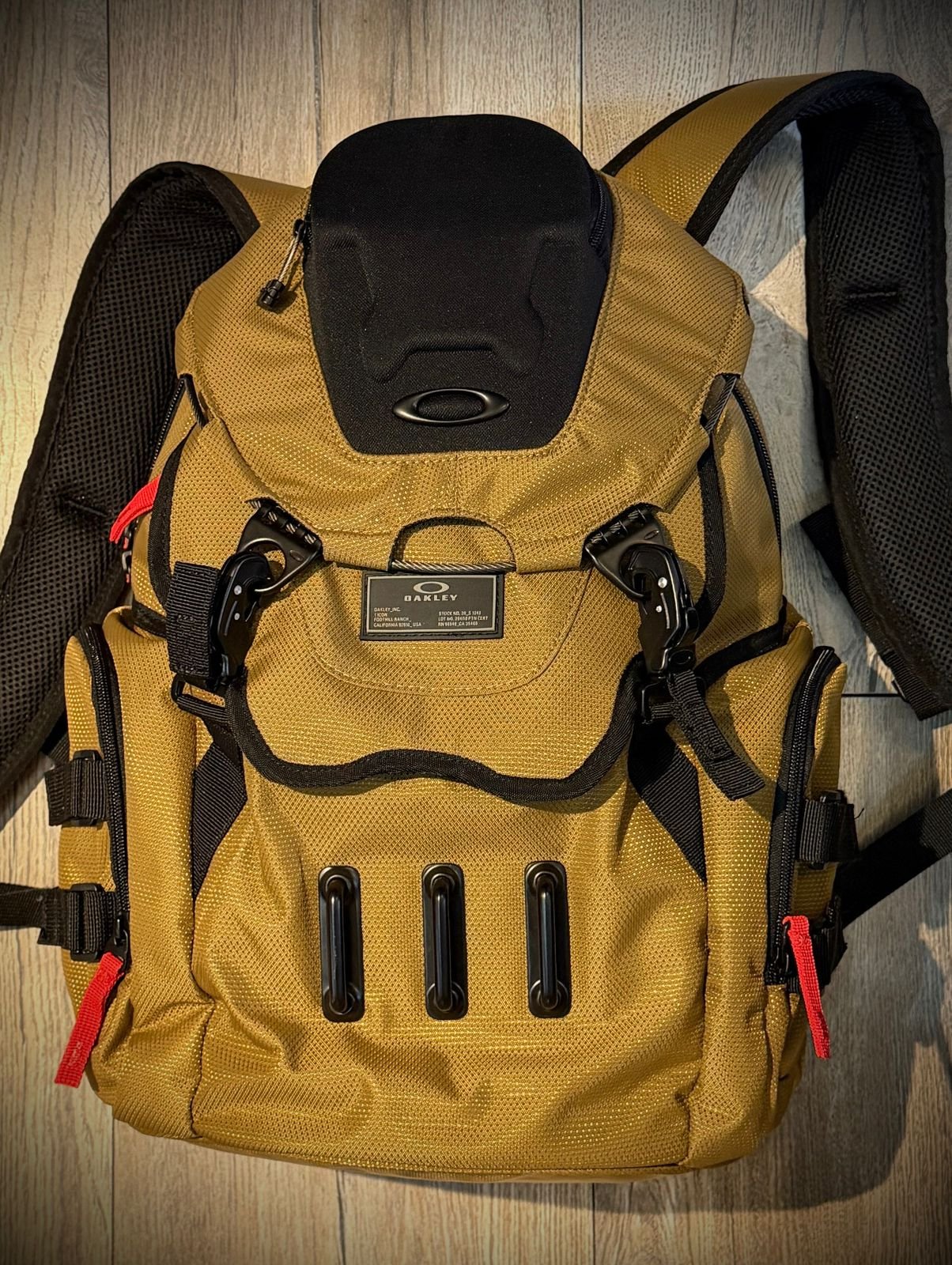 A tan and black Oakley backpack with multiple compartments and straps, placed on a wooden floor.