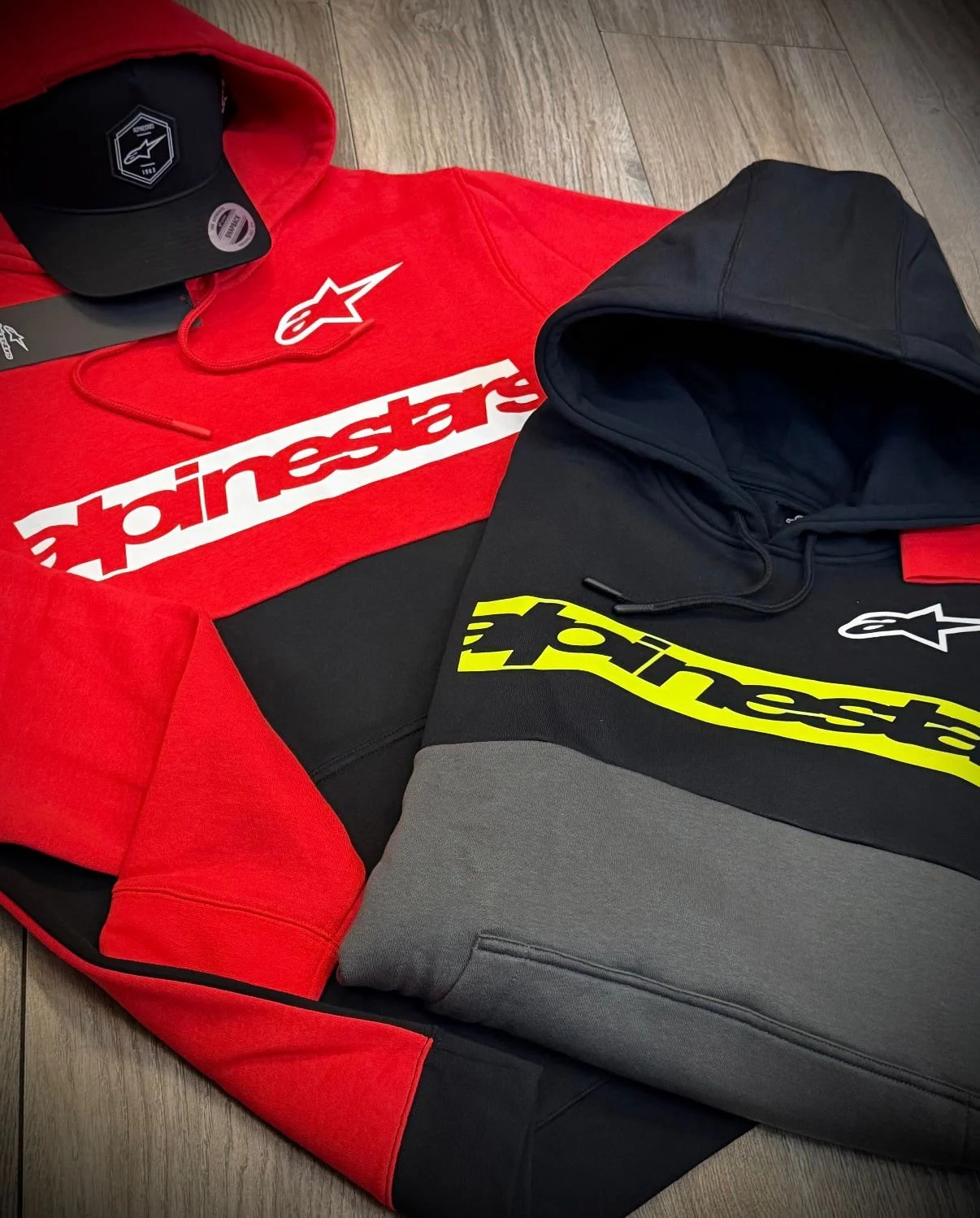 Red and black Alpinestars hoodie, gray sweatpants, black cap with logo, black and yellow Alpinestars hoodie, and black sunglasses on a wooden floor.