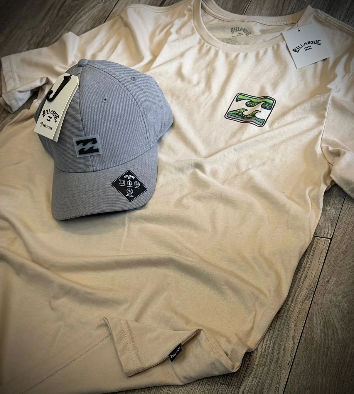 A beige t-shirt and a gray Billabong cap with tags on a wooden floor.