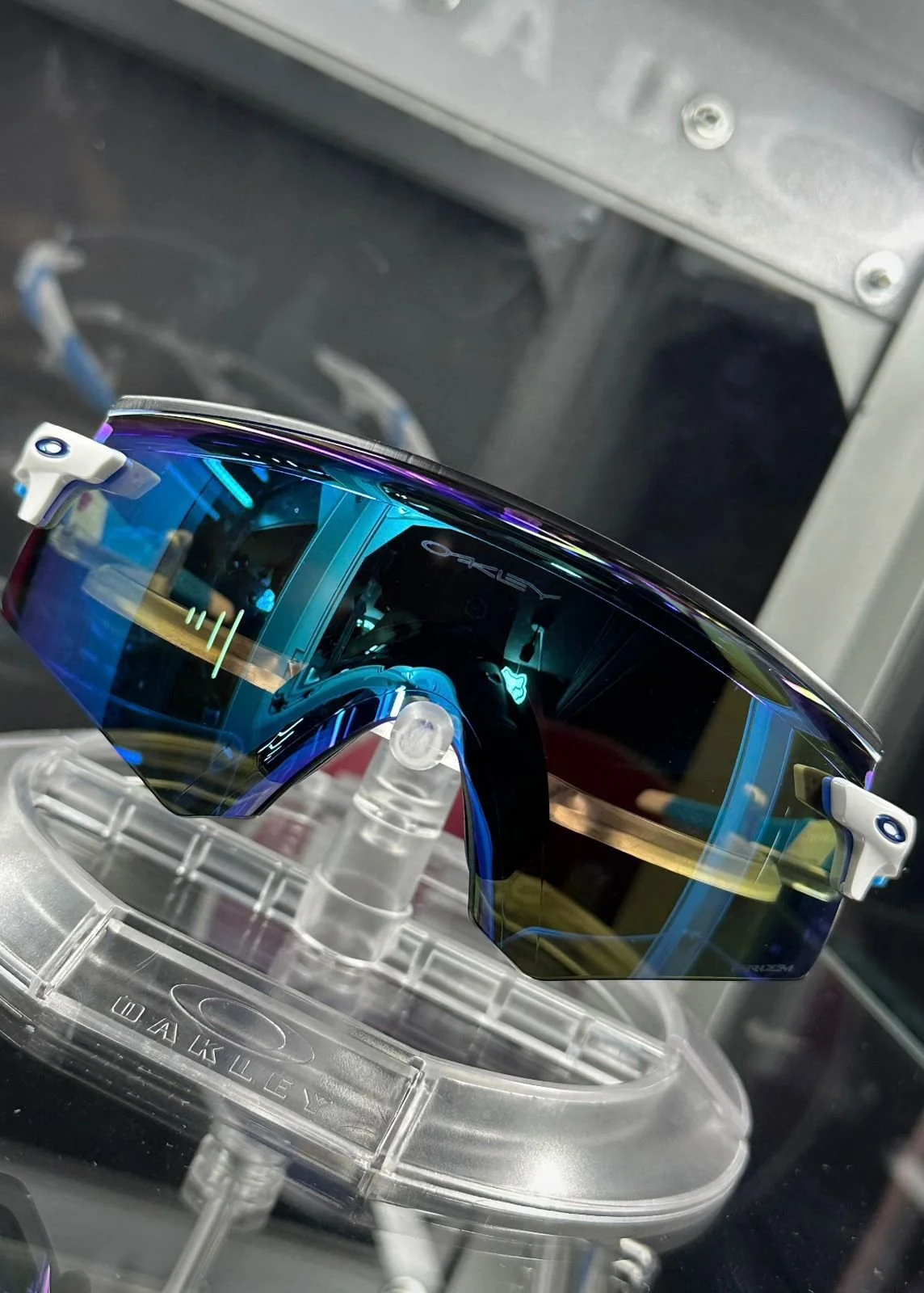 A pair of Oakley sports sunglasses with a mirrored, reflective blue lens displayed on a clear plastic stand.