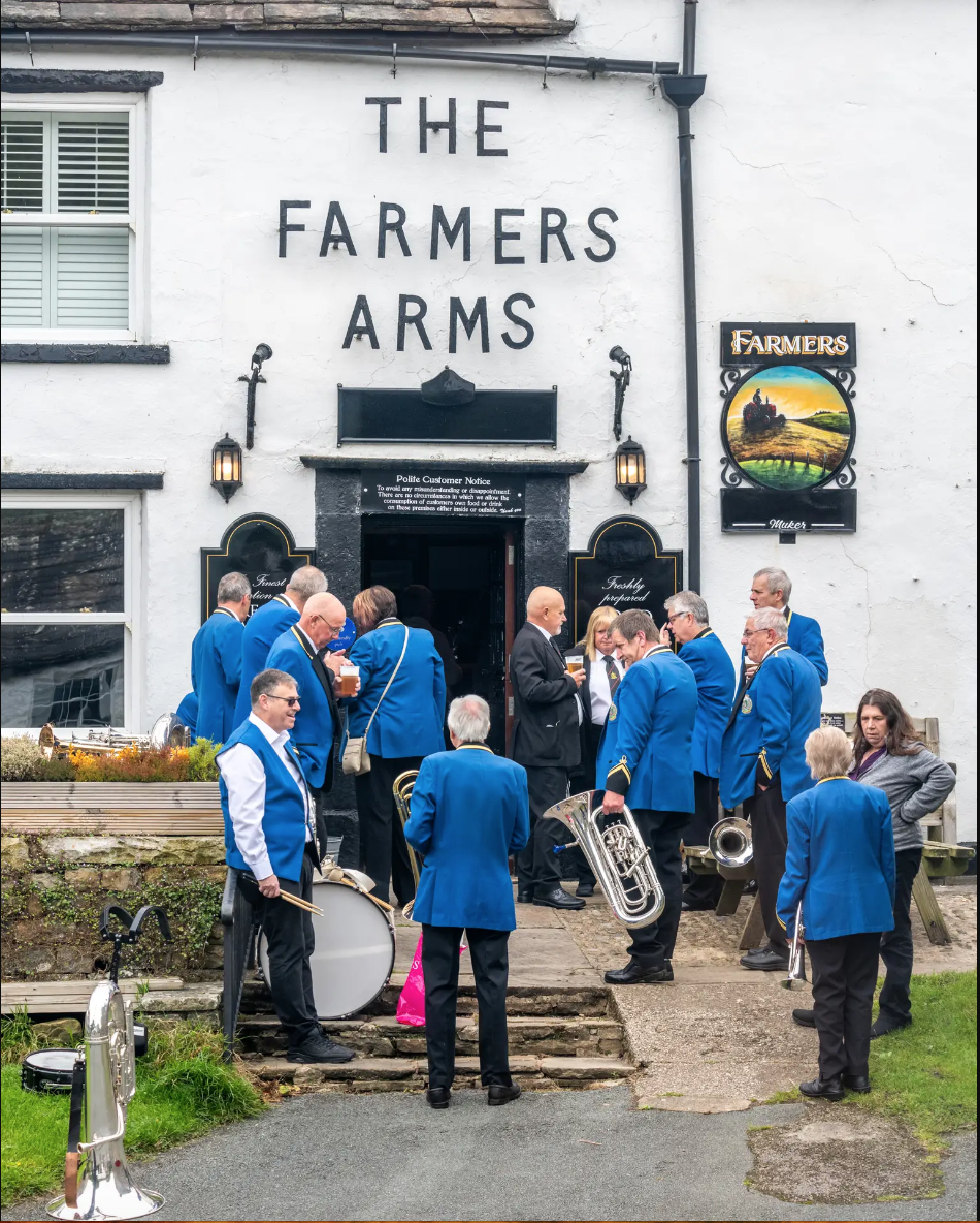 A group of musicians in blue uniforms gathering outside a pub called The Farmers Arms, with some holding musical instruments and drinks, on a patio in front of a white building.