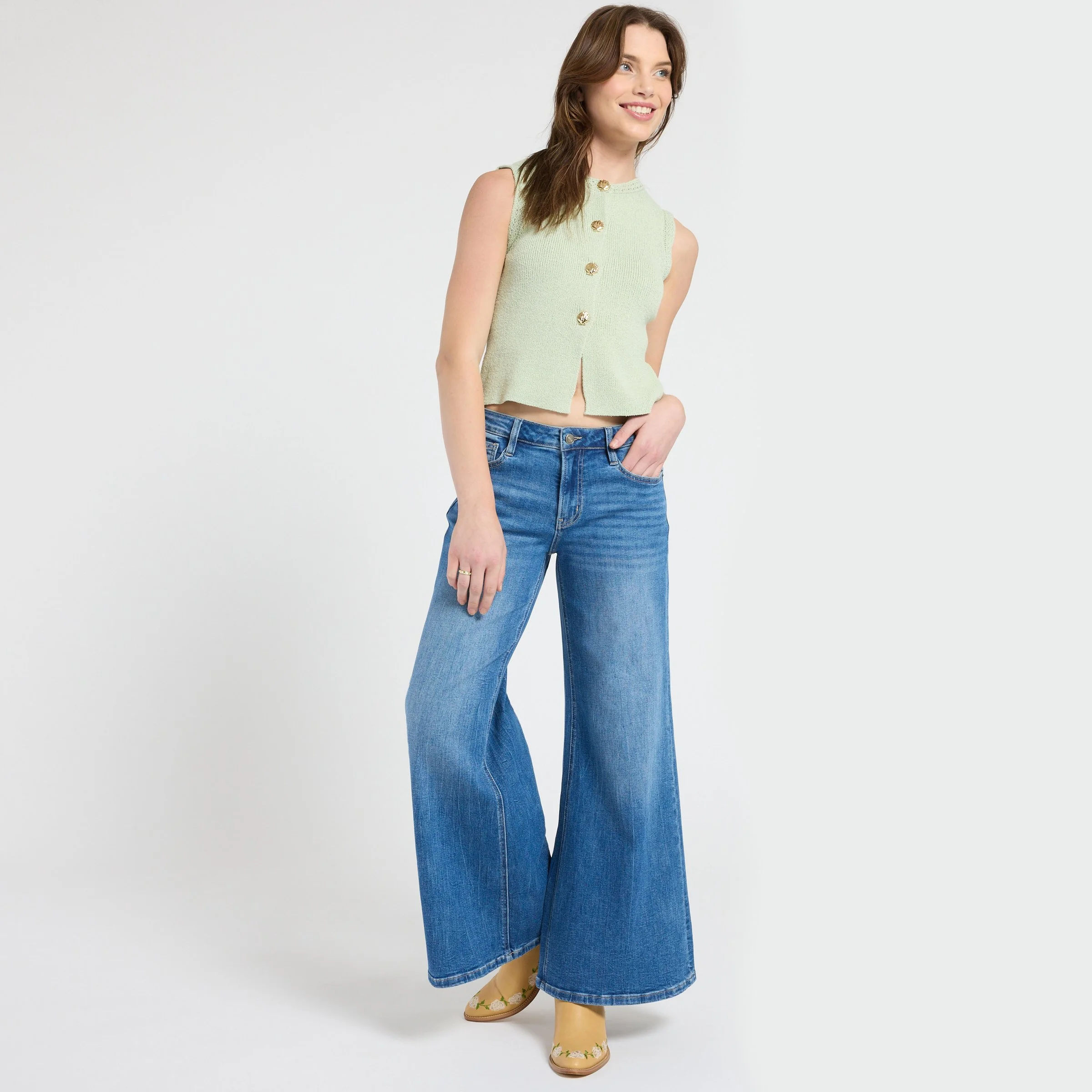 Smiling woman modeling a mint green sleeveless button-front sweater top, wide-leg blue jeans, and floral shoes against a white background