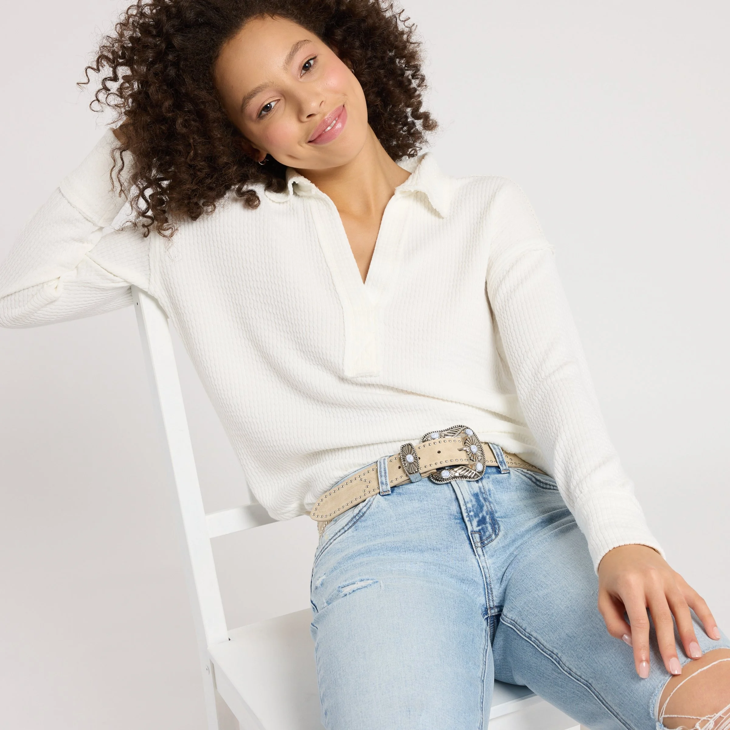 Smiling woman with curly hair wearing a white textured blouse, light blue jeans, and a decorative belt while sitting on a white chair.