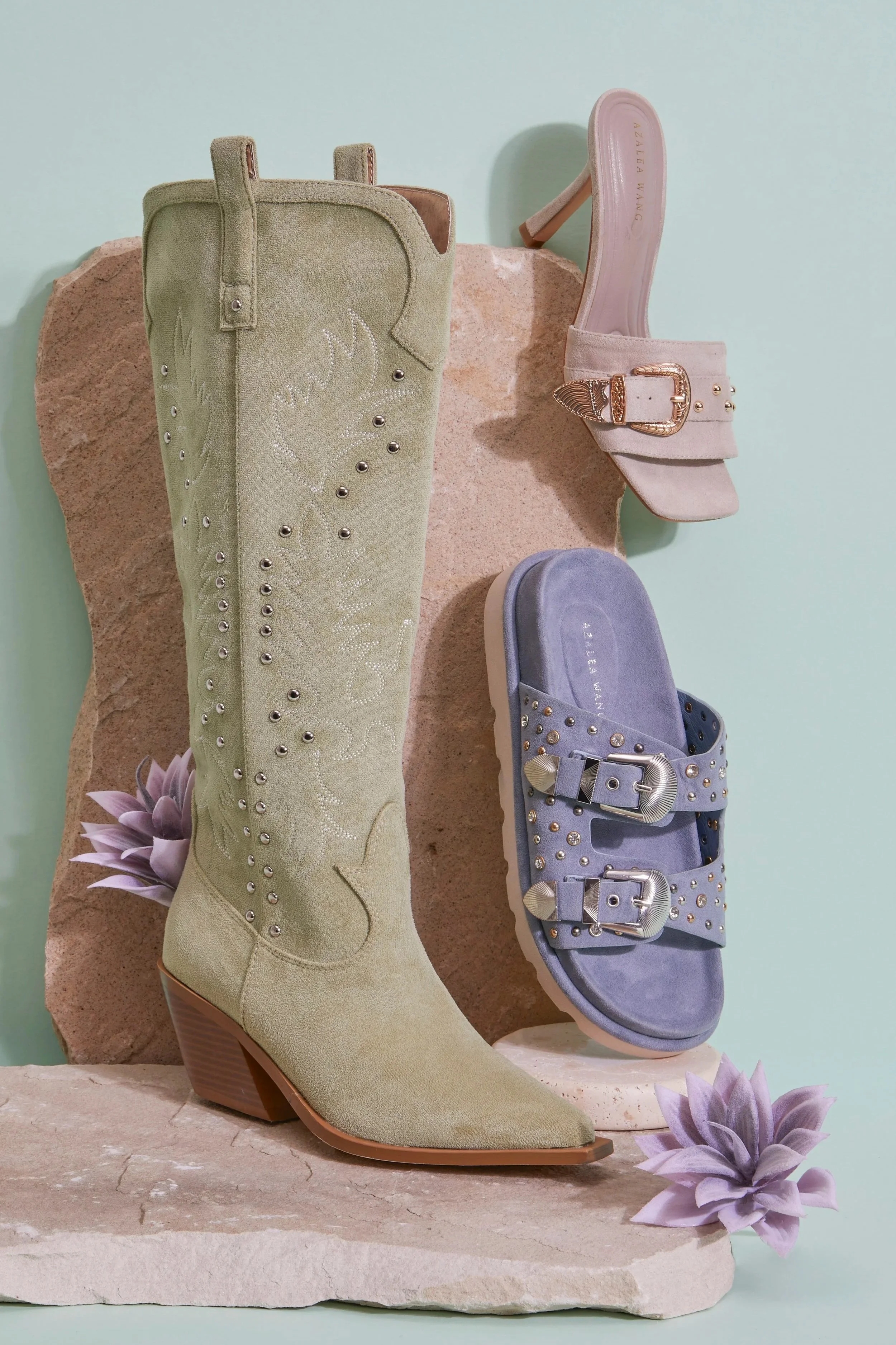 Green studded pointed-toe ankle boot and lavender buckled sandals displayed on a stone pedestal with succulents