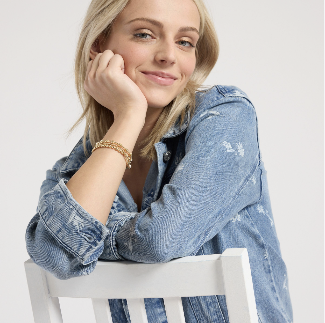 Smiling blonde woman wearing a floral denim jacket and gold bracelets, resting her chin on her hand behind a white chair