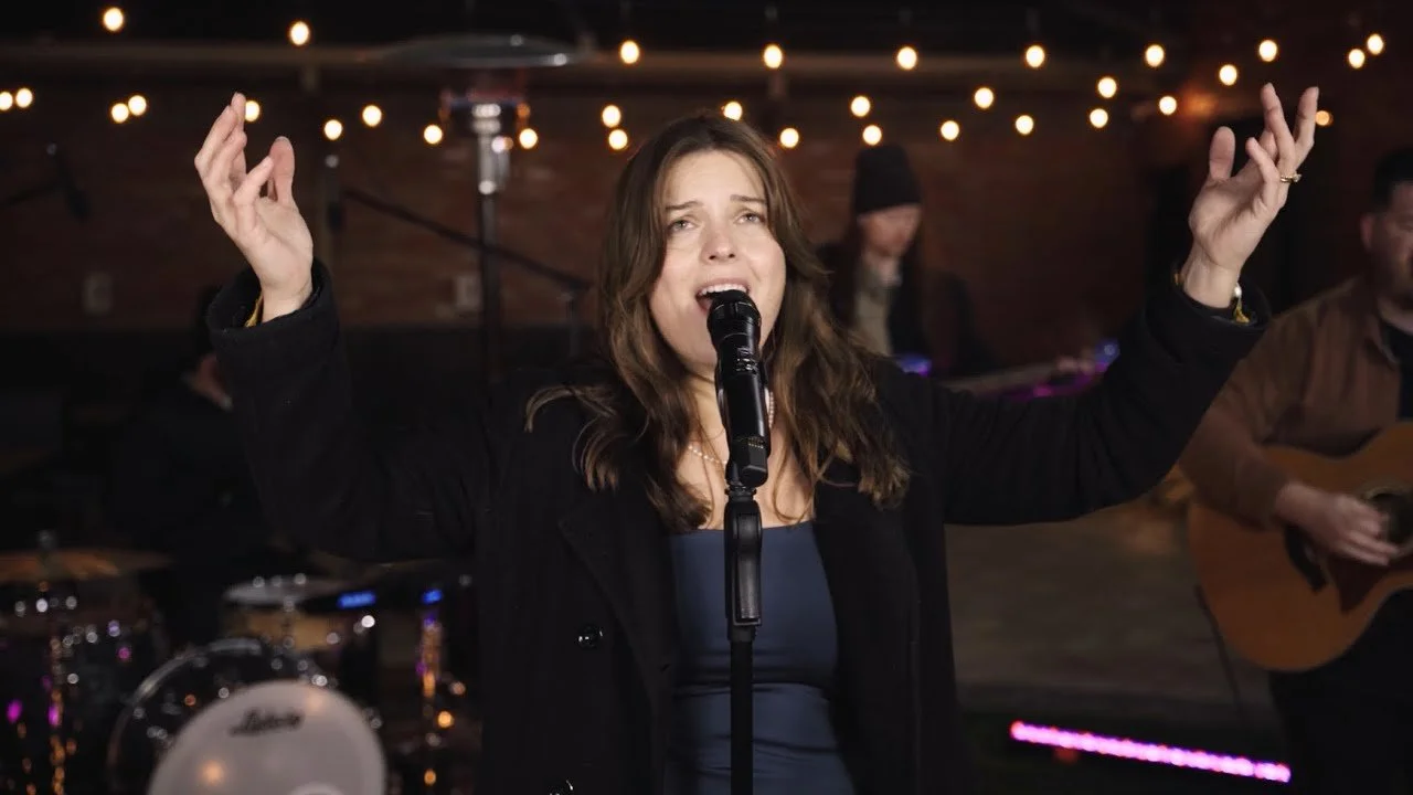 A woman singing into a microphone during a performance, with a band in the background, string lights overhead, and a stage setting.
