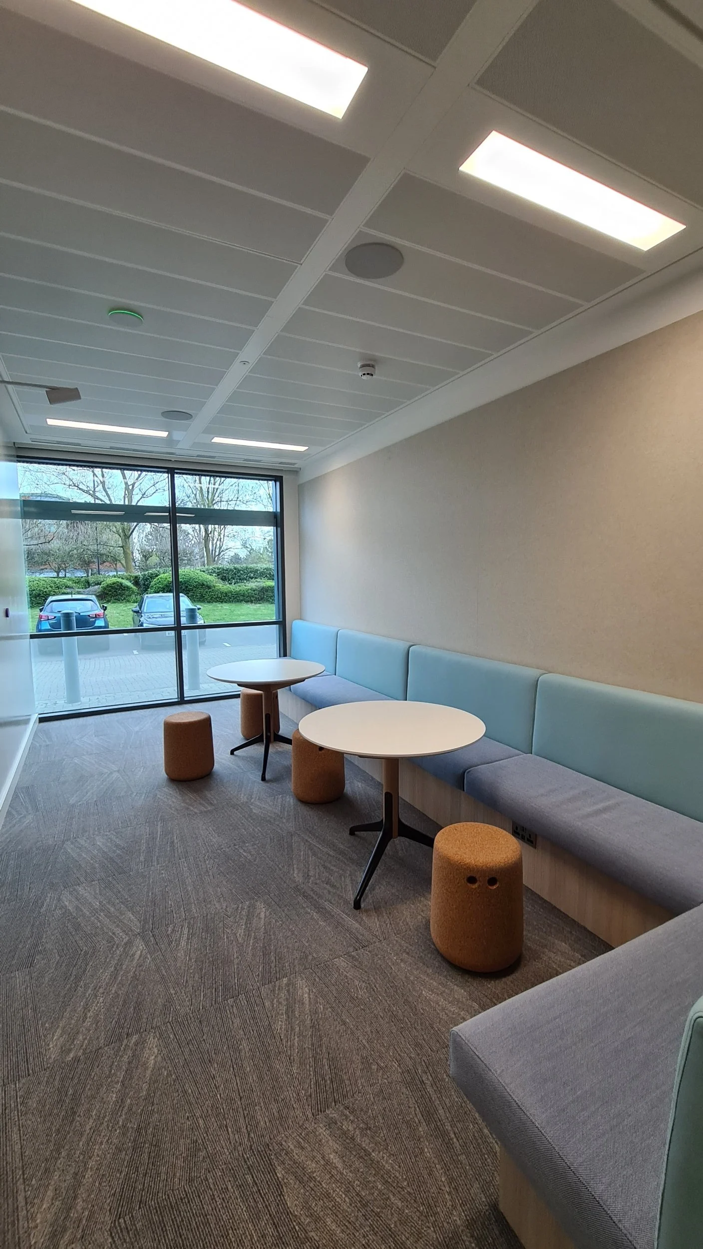 Empty lounge area with light blue and gray seating, round white tables, orange ottomans, large glass window showing parked cars and trees outside.
