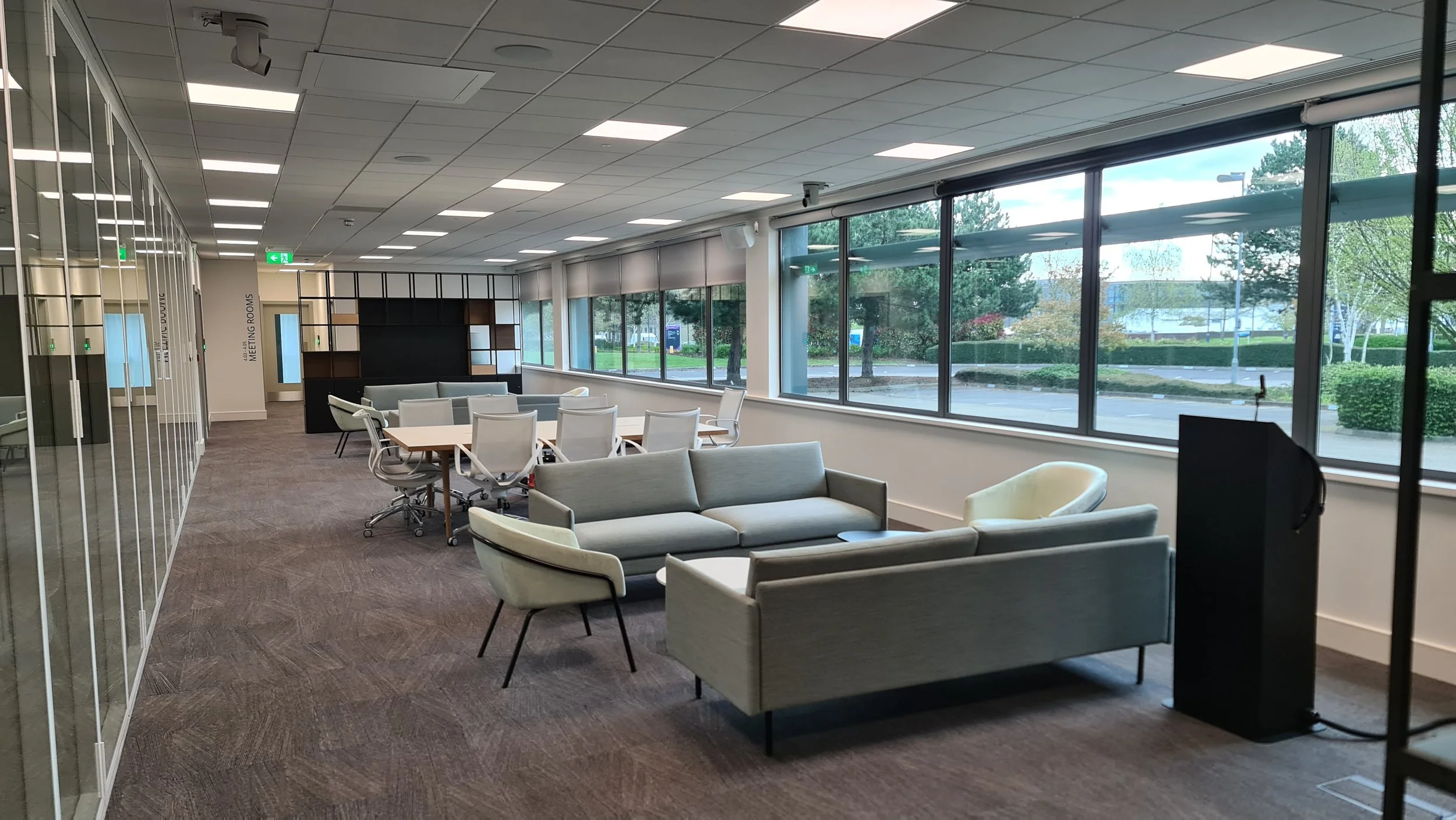 Modern office lounge with large windows, white sofas and chairs, a conference table with white chairs, and a glass meeting room with frosted doors.