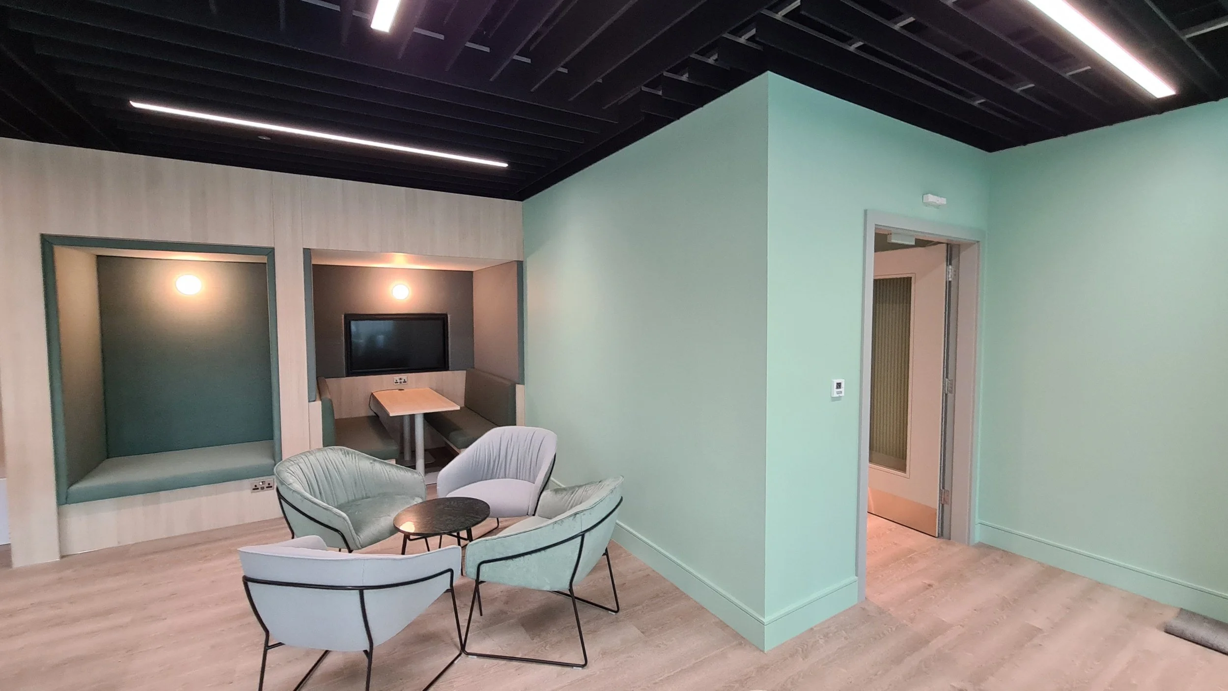 Modern lounge area with pastel green walls, light wood flooring, and contemporary furniture, including a round table, armchairs, and built-in seating alcoves with a TV and lighting.