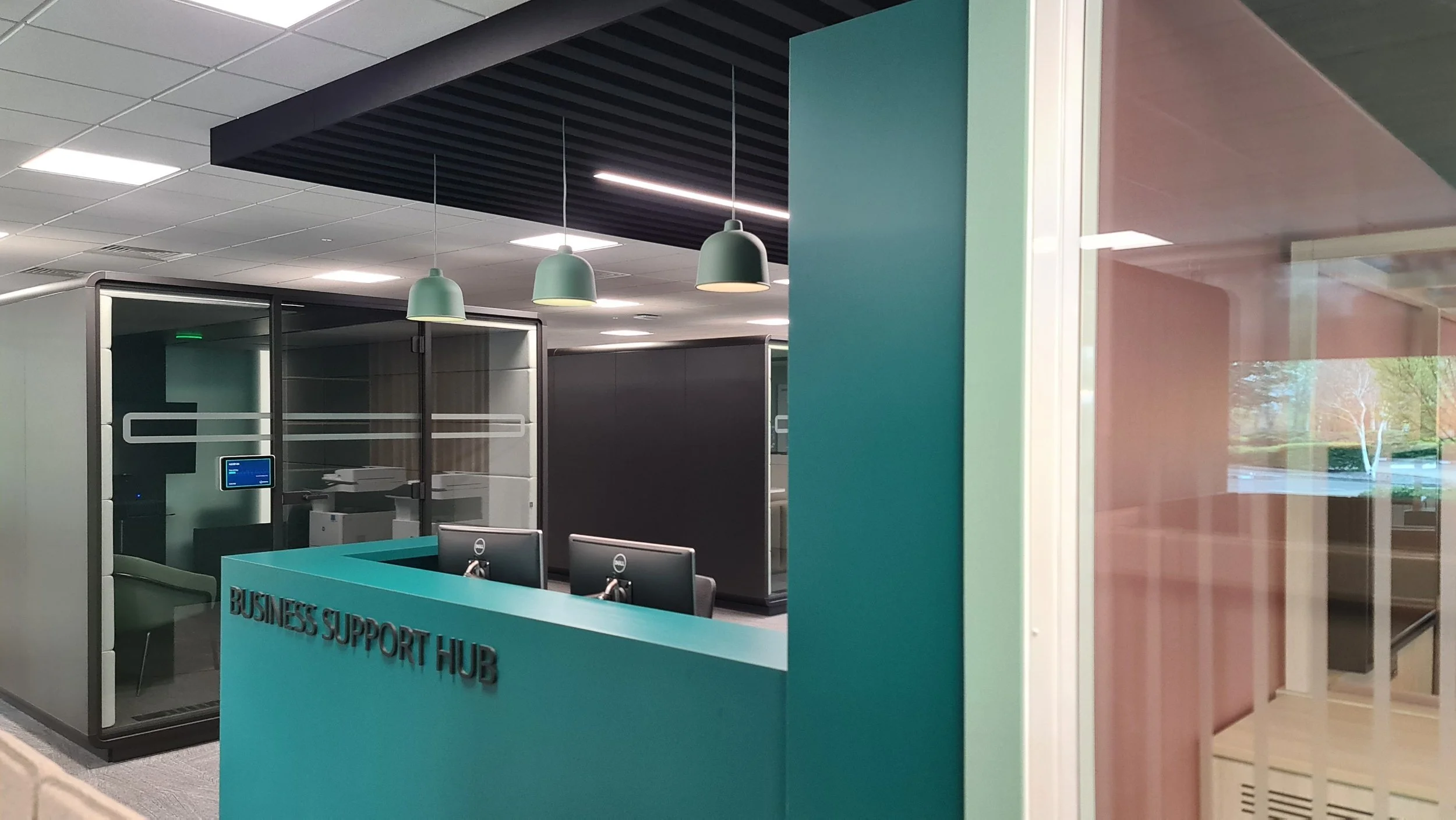 Reception desk with computers at a business support hub, glass-enclosed workspace, teal and pink walls, hanging teal lamps, and a view of trees outside through a window.