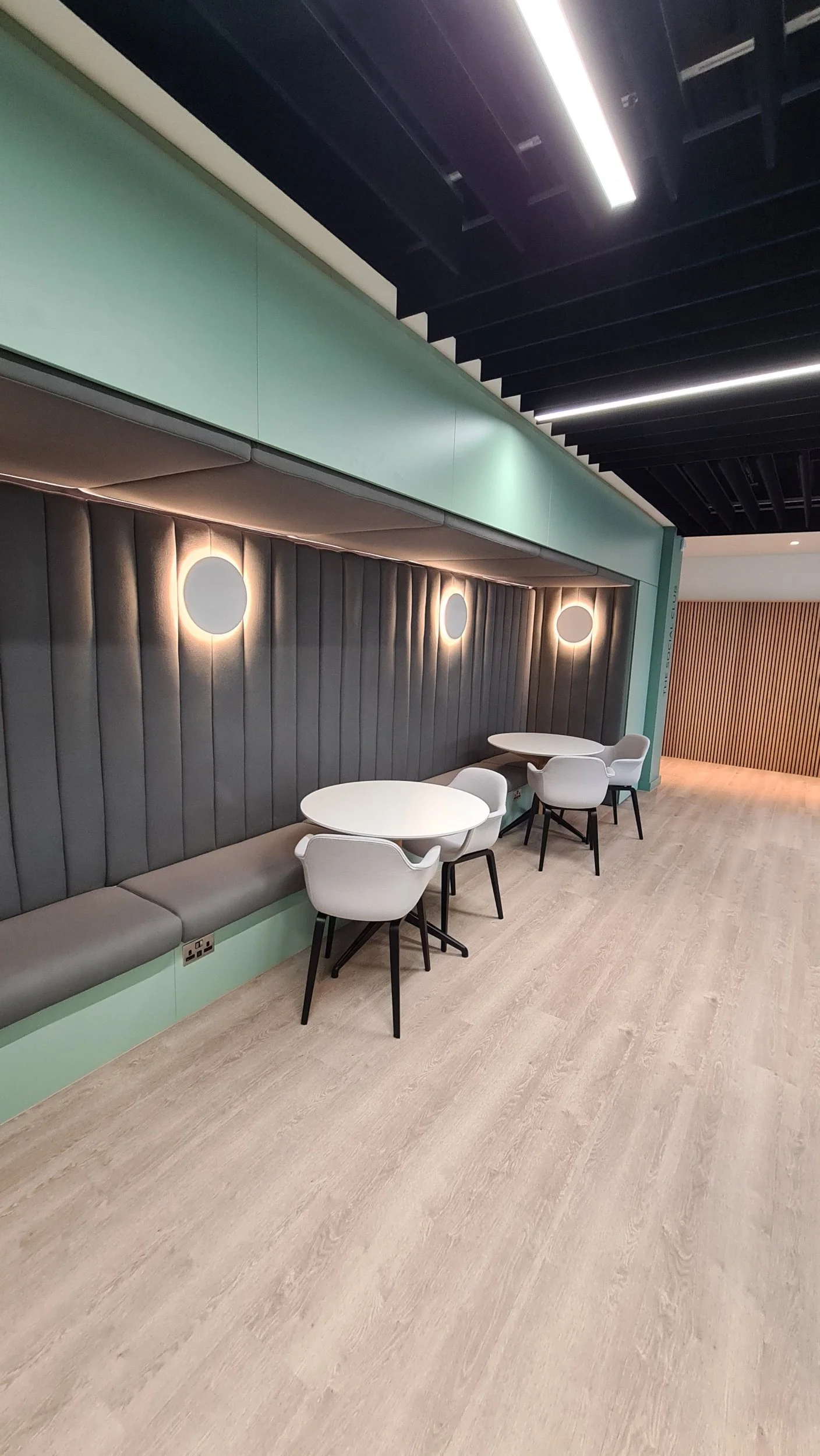 Modern waiting area with three white round tables, white chairs with black legs, a gray padded bench, black ceiling, and decorative wall lights.