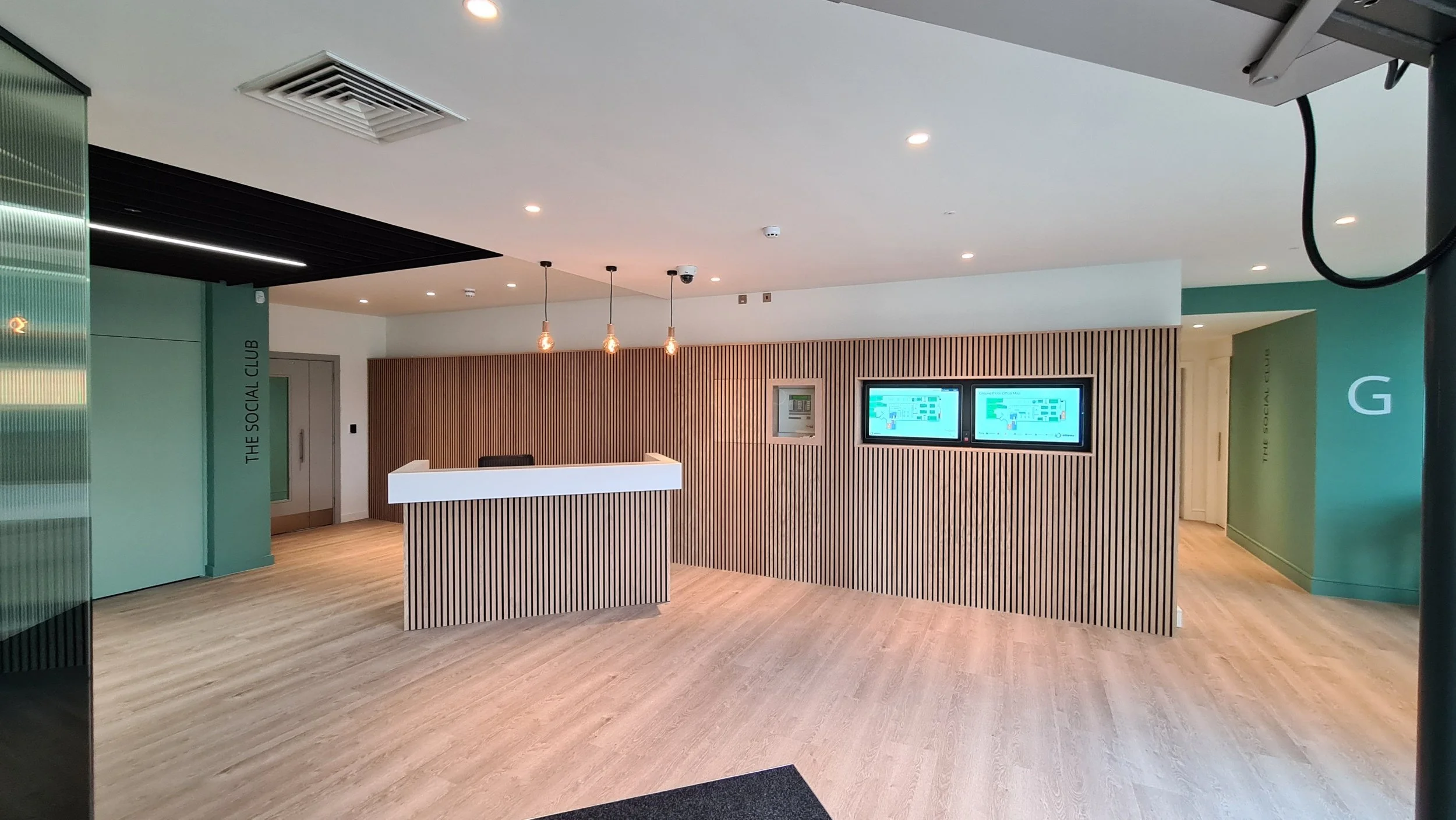 Modern lobby area with a wooden slat wall, reception desk, two digital display screens, and hanging pendant lights.