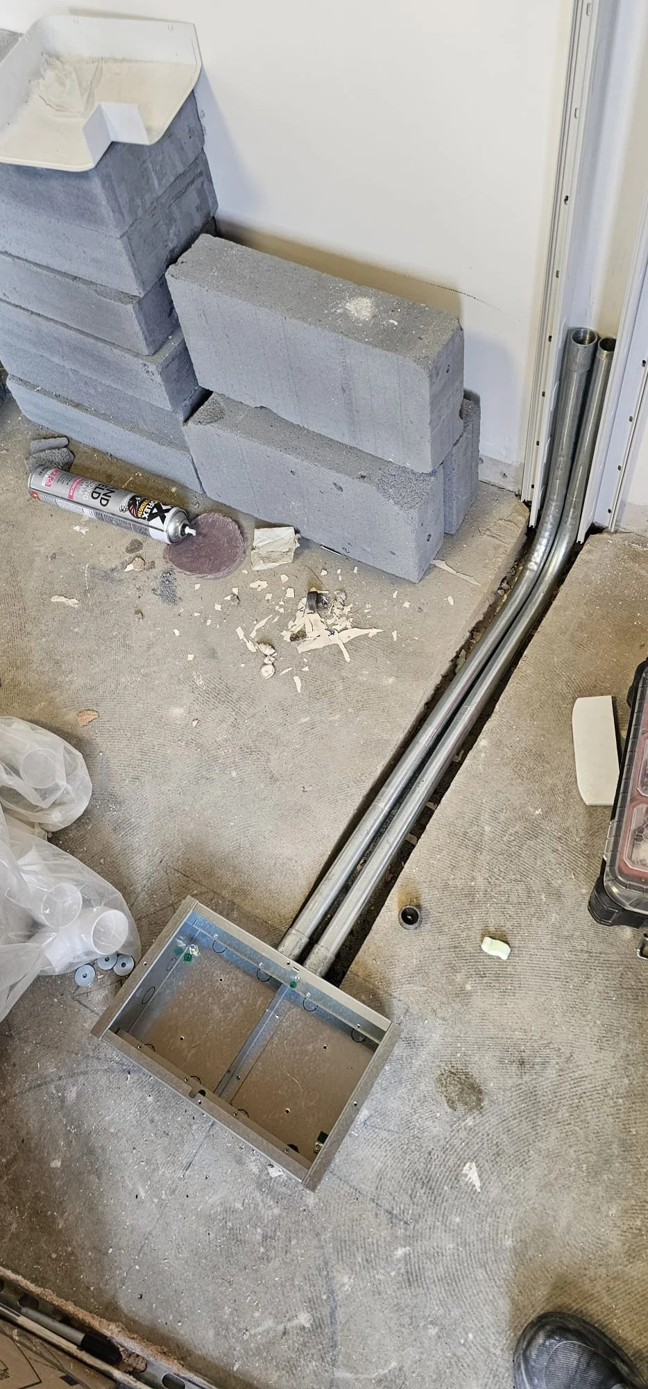 Construction site with concrete blocks, electrical conduit pipes, and a metal box on the floor, surrounded by construction debris and tools.