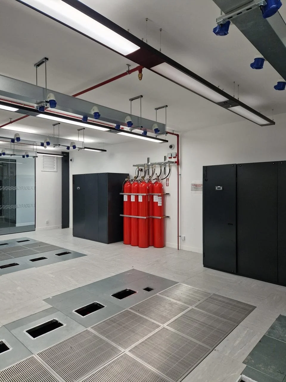 Interior of a modern building with fire safety equipment including red fire extinguishers on the wall, black cabinets, overhead lighting, and floor drains.