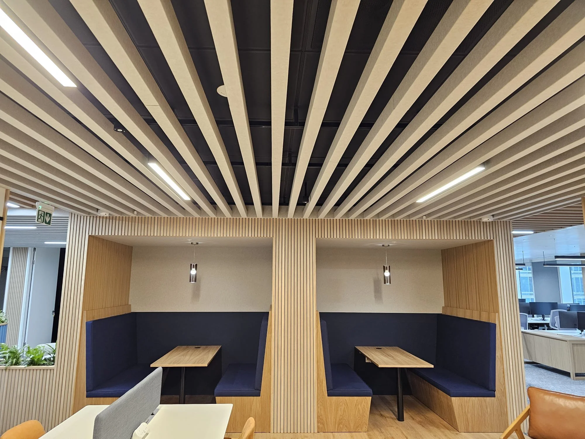 Modern office interior with two booth seating areas featuring dark blue cushioned walls and wooden tables, separated by vertical wooden slats, with a ceiling of parallel wooden slats and integrated lighting.