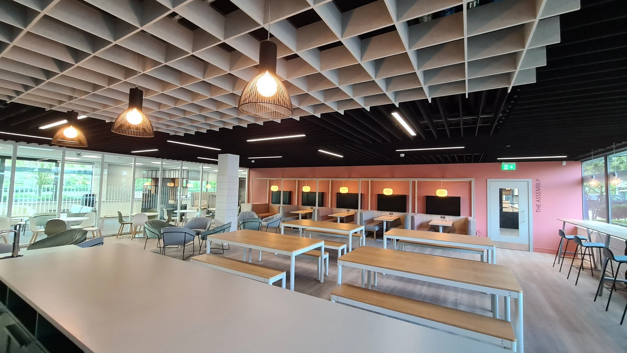 Modern cafeteria with wooden tables, armchairs, and booth seating, large windows, hanging light fixtures, and a decorative ceiling.
