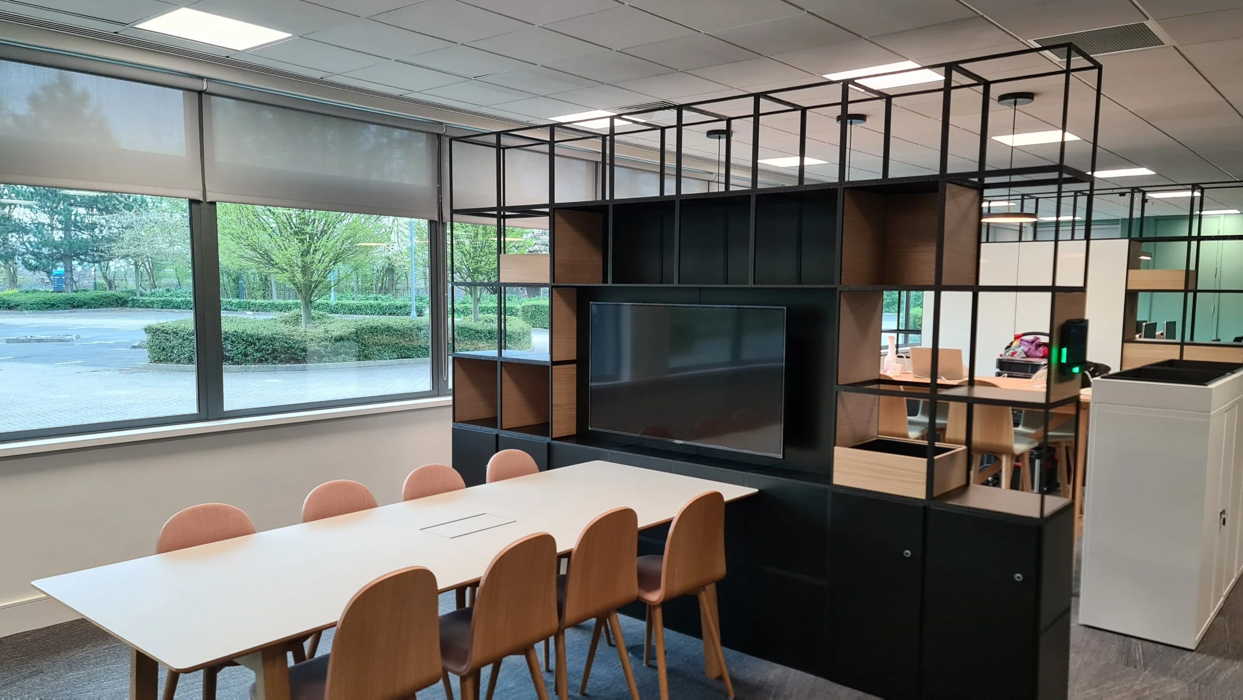 Modern office conference room with a white table, six pink chairs, and a large black shelving unit with a TV, located by large windows showing a green outdoor landscape.