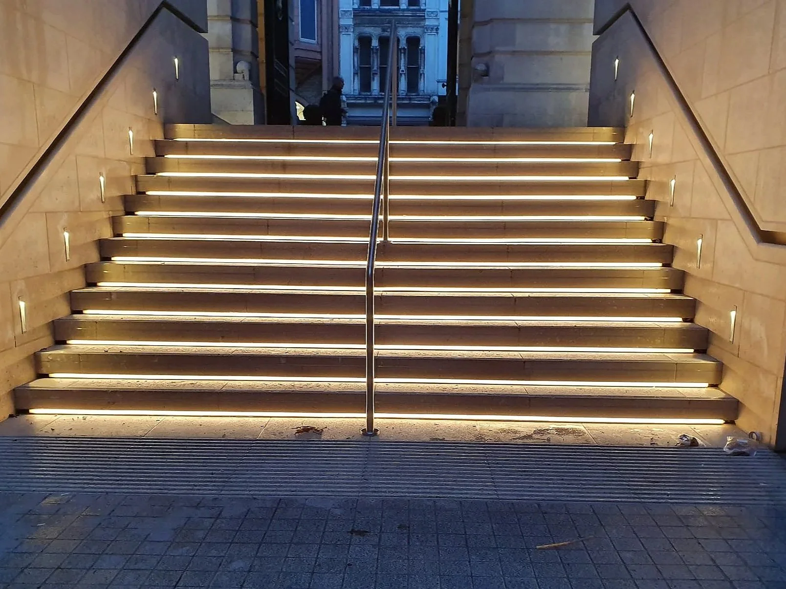 Exterior staircase with illuminated steps leading up to a building entrance, with decorative lighting on side walls and a metal handrail down the center.