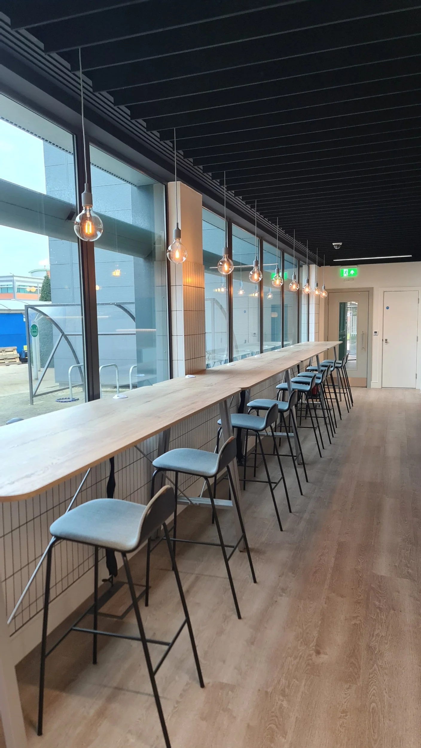 Empty modern cafe with a long wooden counter, chairs, and hanging light bulbs near large windows.