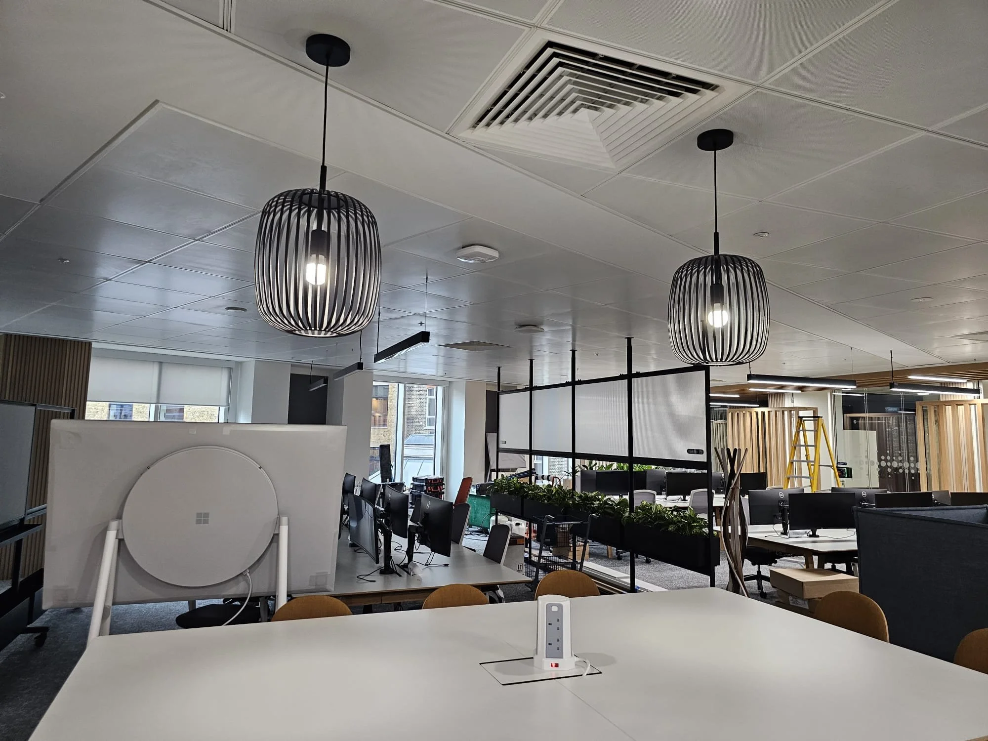 Modern office space with a white table in the foreground, computer monitors, pendant lights, partition walls, a yellow ladder, and various office furniture and decor.