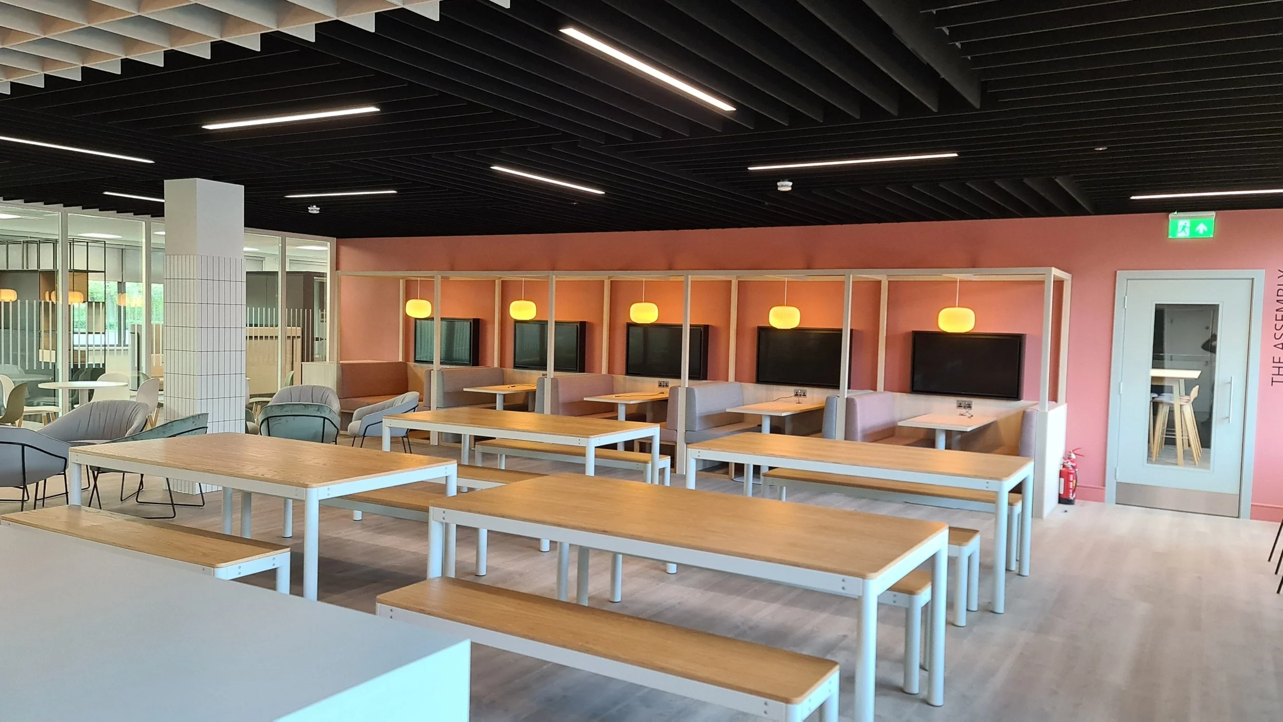 Empty modern cafeteria with wooden tables and benches, booth seating with TVs, hanging lights, and glass partition in the background.