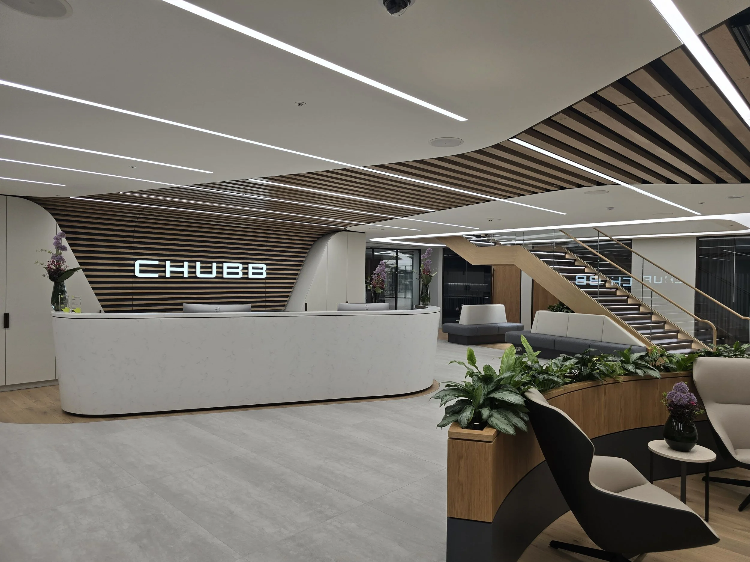 Modern office lobby with white reception desk, wooden accents, black and white seating, and green plants, with the sign 'CHUBB' on the wall.