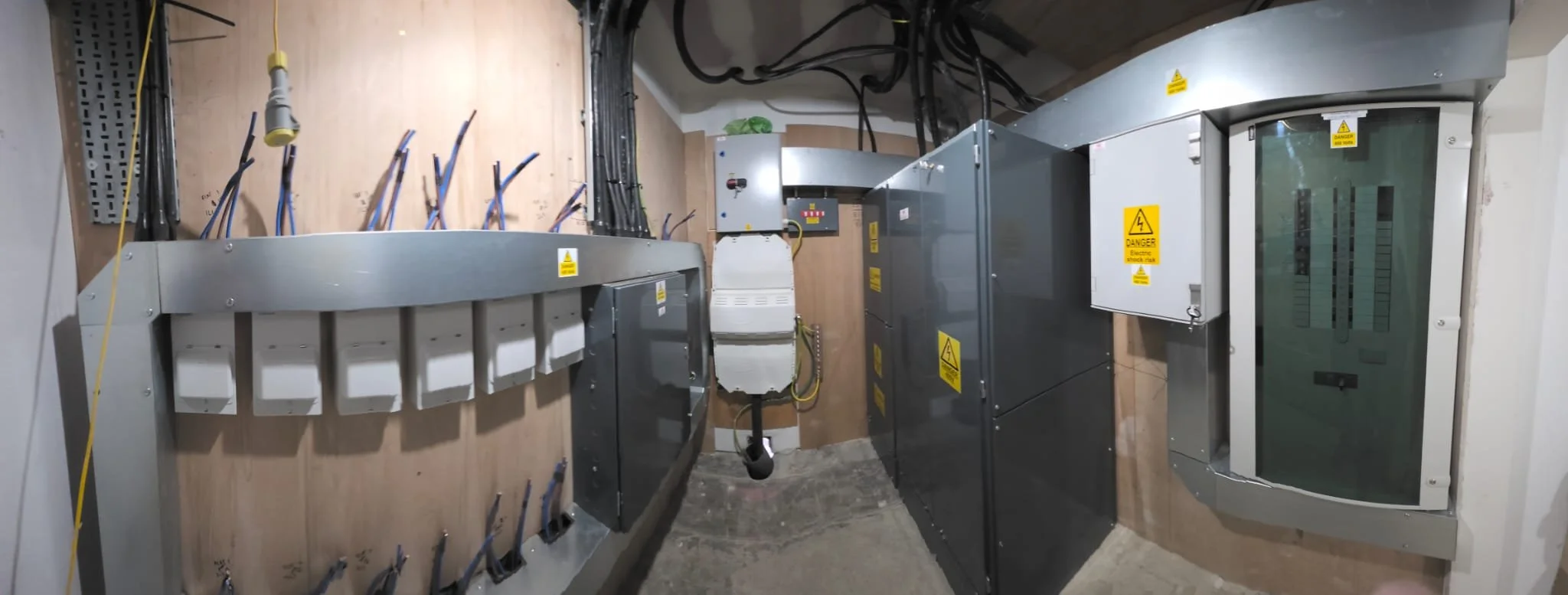 Electrical panel room with circuit breakers and wiring enclosed in metal cabinets; several warning labels indicating electrical hazards.