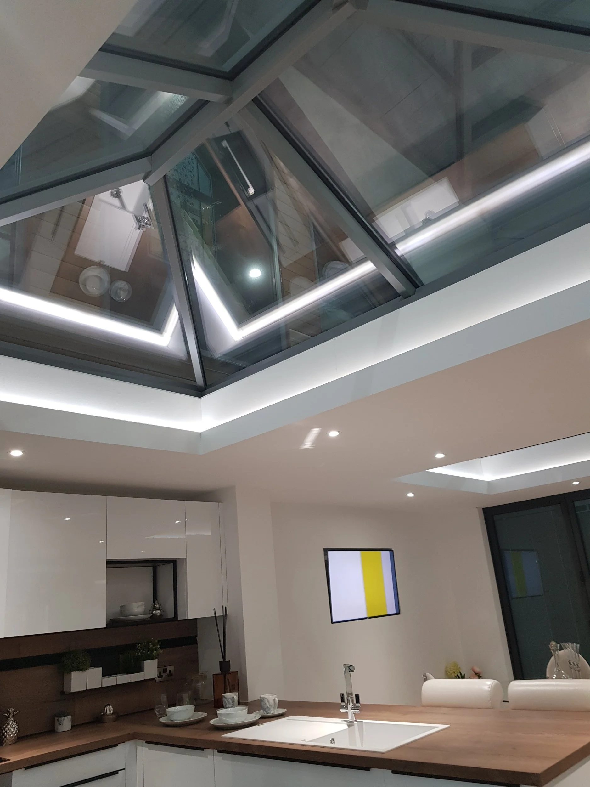 Modern kitchen with white cabinets, wooden countertop, and a sink. A small wall-mounted TV displays a yellow and white pattern. The ceiling features a skylight with multiple glass panels and white LED lighting.