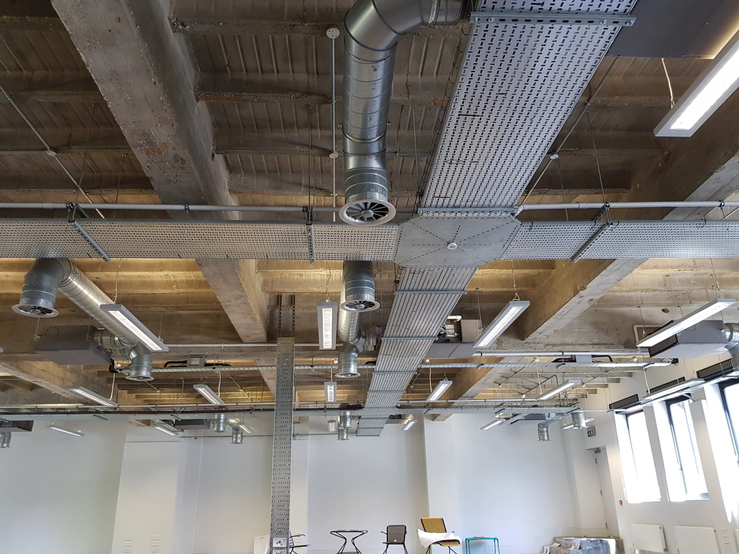 Ceiling of an industrial-style interior with exposed concrete beams, metal ducts, perforated metal panels, suspended rectangular LED lights, and large windows letting in natural light