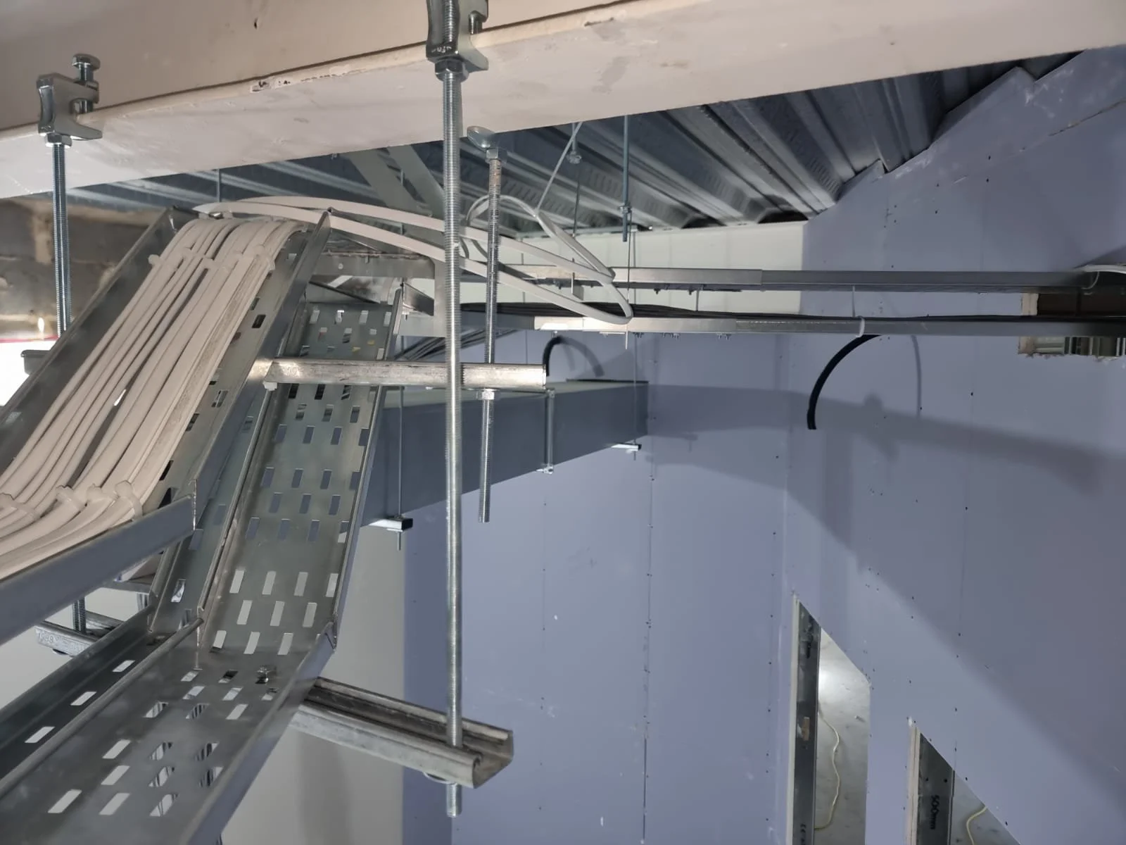 Interior view of a construction site showing electrical conduit and cable management systems installed on a metal framework attached to the ceiling and wall.