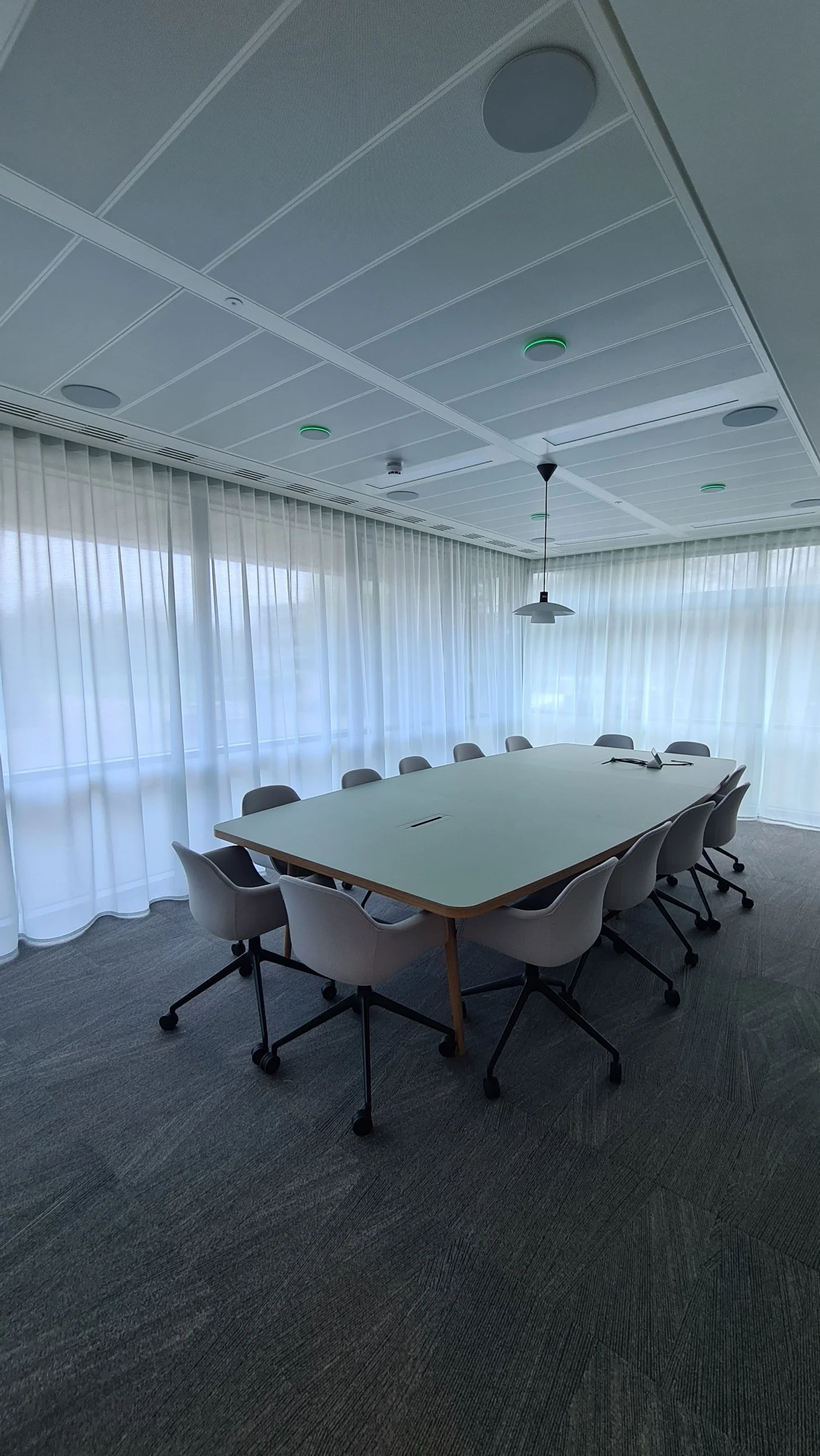 Empty conference room with a large white table, ten gray chairs, a projector screen on the table, and large windows with sheer curtains.
