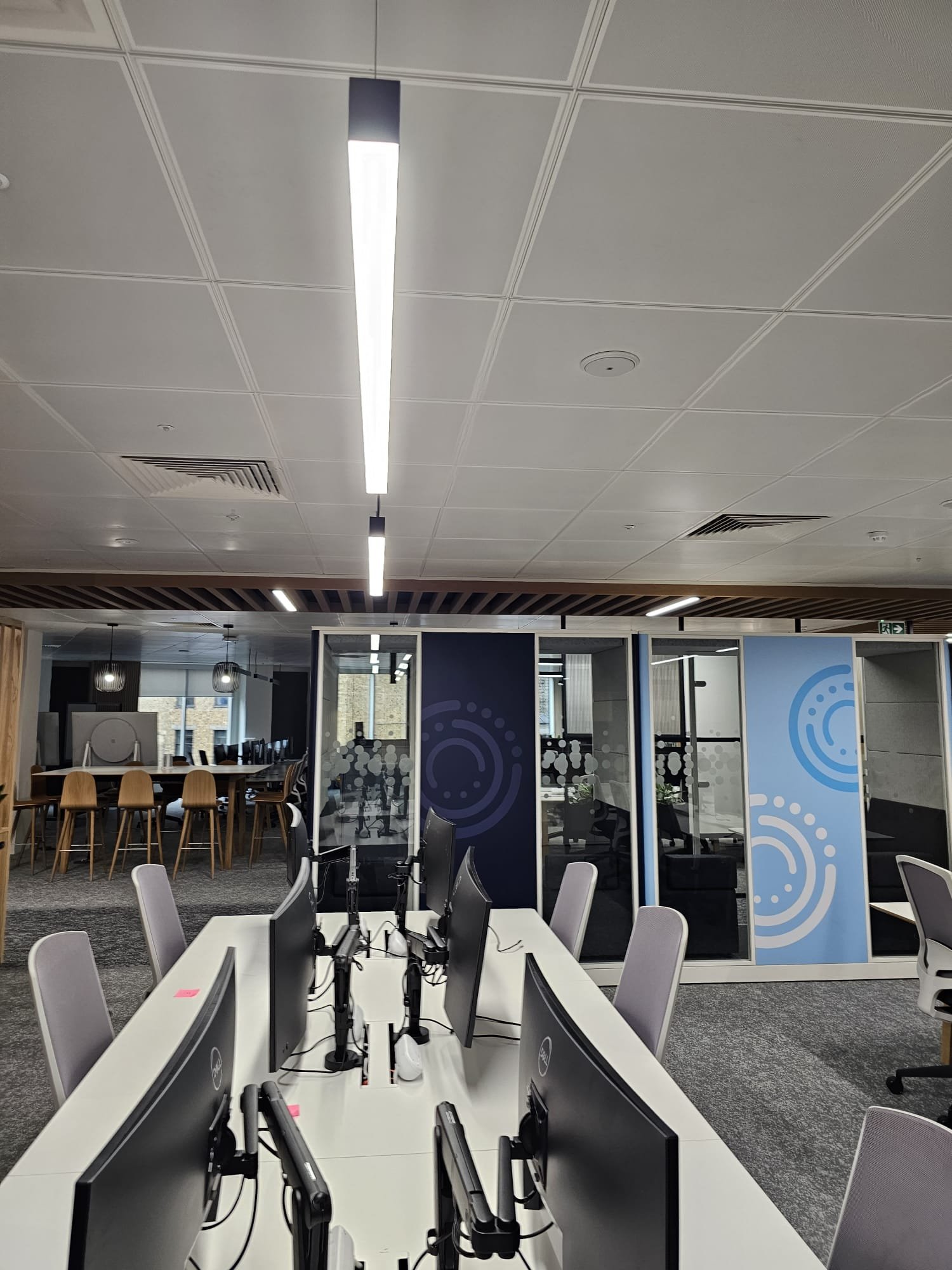 Modern office workspace with multiple computer monitors on a white desk, surrounded by gray chairs, in an open-plan office with partitioned meeting rooms featuring glass walls with circular patterns, and a background with tables and windows.