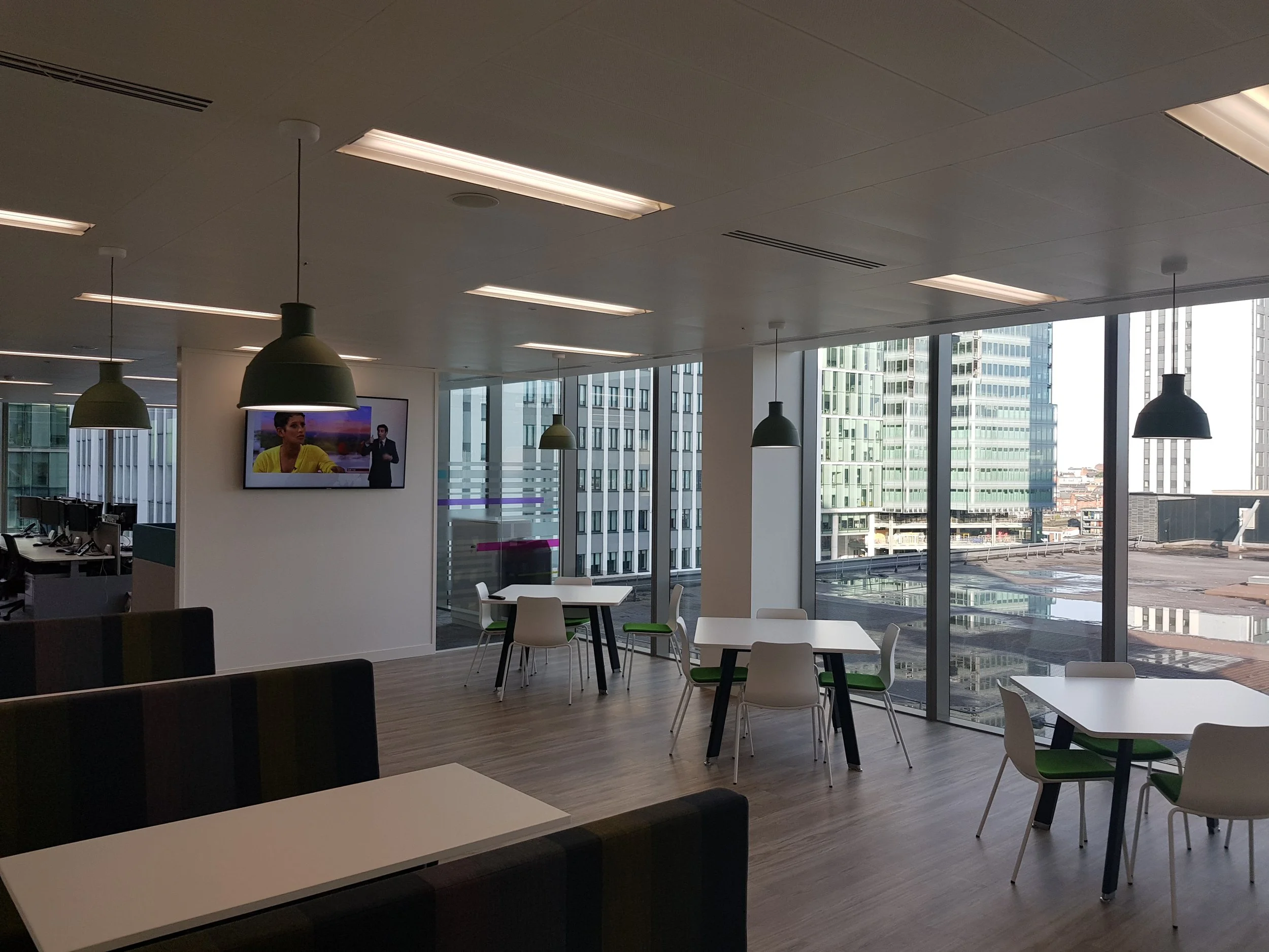 Modern office break room with white tables, white chairs with green cushions, large windows showing city buildings, a TV on the wall, and hanging pendant lights.
