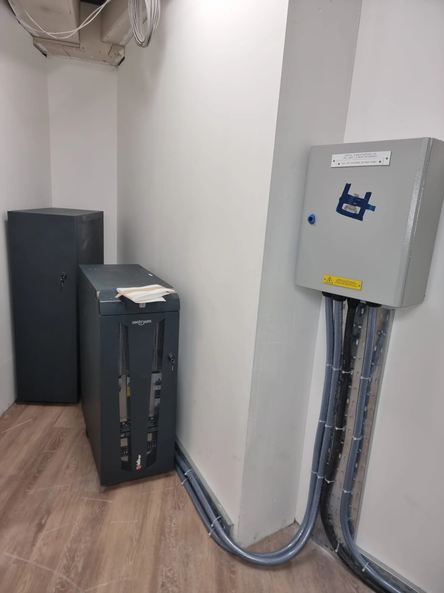 Electrical equipment including a gray electrical panel and a gray UPS system in a utility room with wooden flooring.