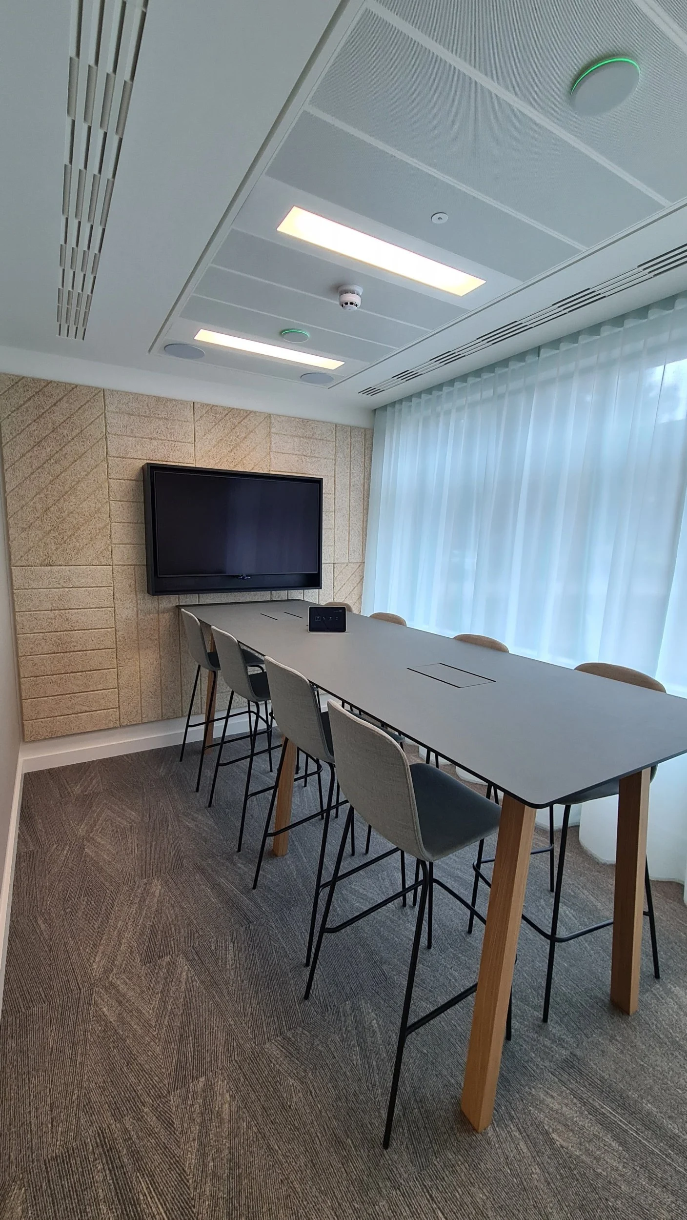 Modern conference room with high table, six chairs, large window with white curtains, flat-screen TV on wall, ceiling lights, and beige textured wall.