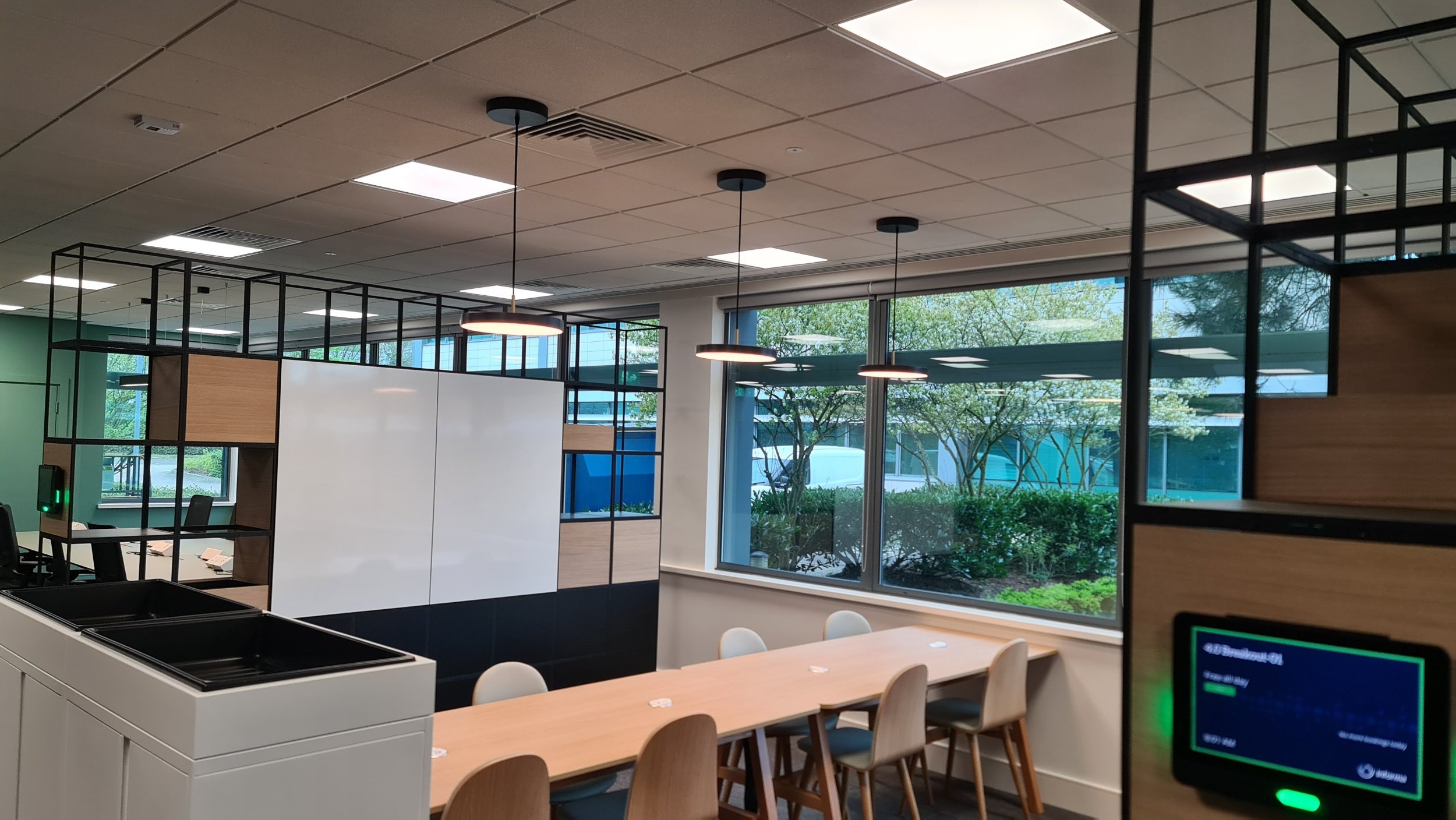 Modern office meeting room with a long wooden table, white chairs, large windows showing greenery outside, black and wooden shelving, and ceiling lights.