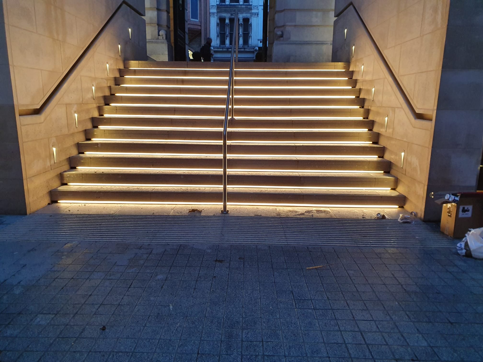 Outdoor staircase with illuminated steps, metal handrail in the center, flanked by beige stone walls, leading up to a building entrance at dusk.