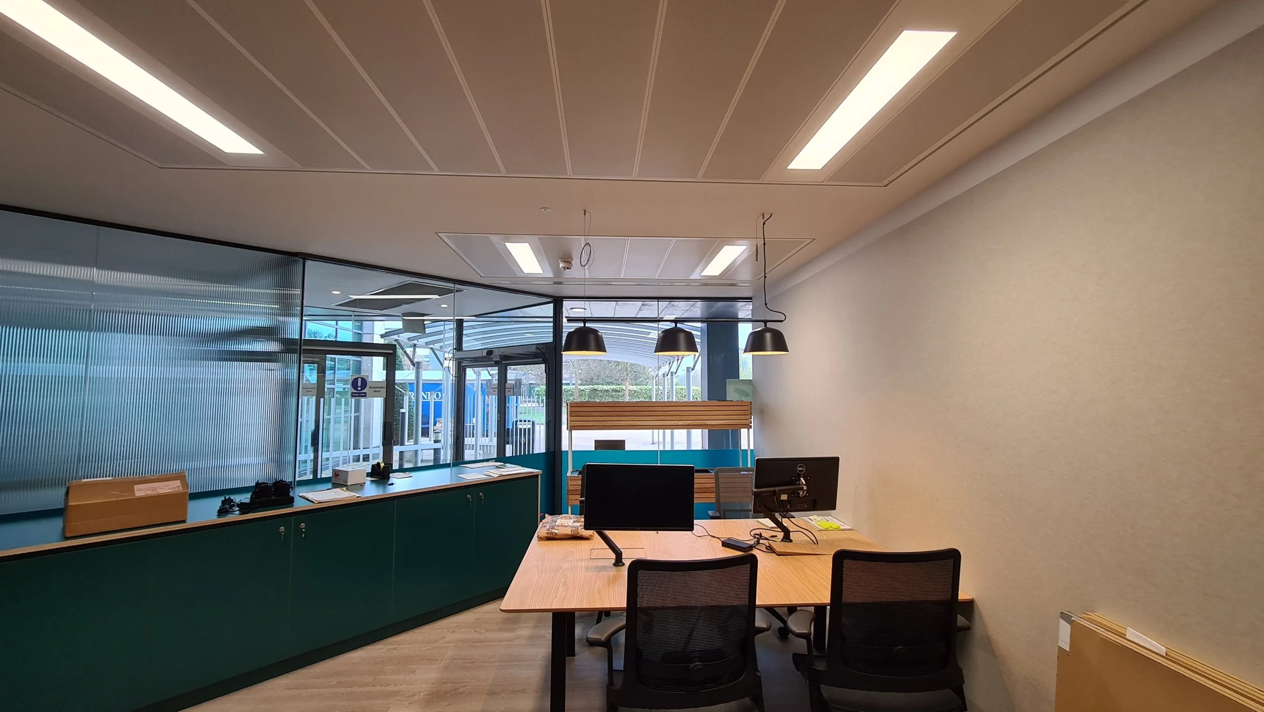 Empty office reception area with two computer desks, three chairs, hanging lamps, and a glass entrance door.