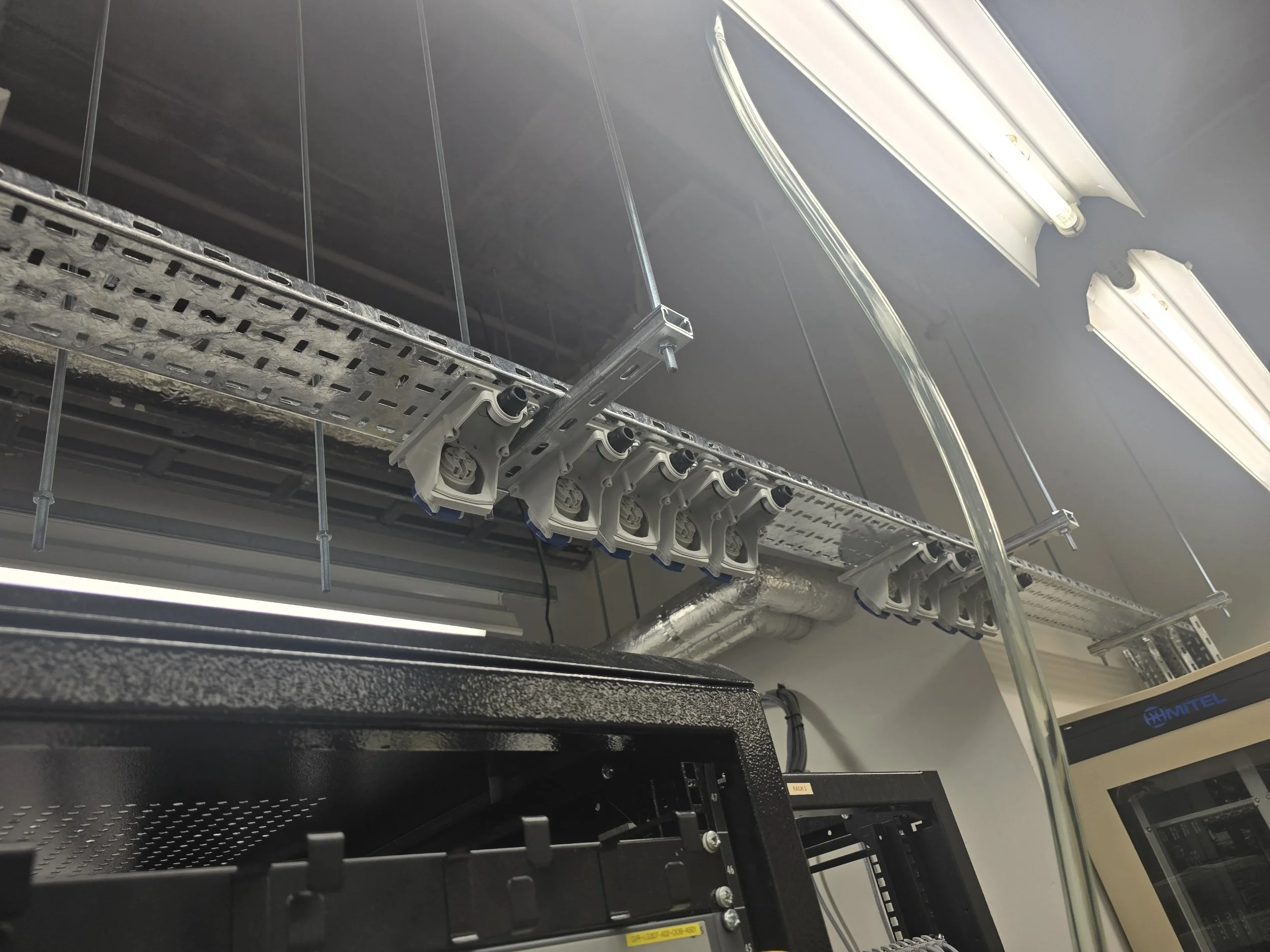 Server rack with cable management and cable conduits in a data center or server room, illuminated by fluorescent ceiling lights.