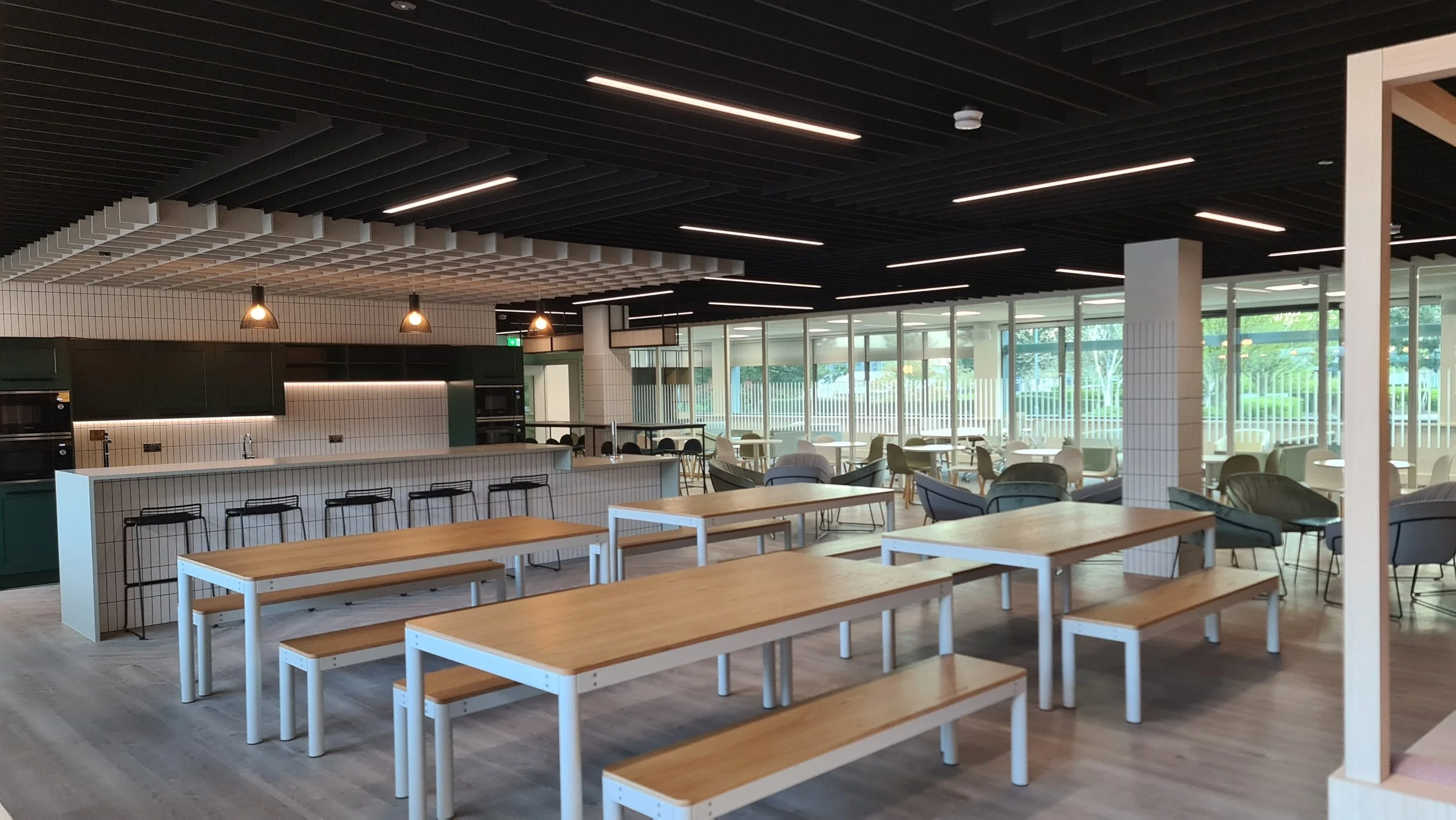 Modern, empty café or lounge with wooden tables and benches, bar with black stools, chairs, armchairs, large windows with outdoor view, and ceiling lights.