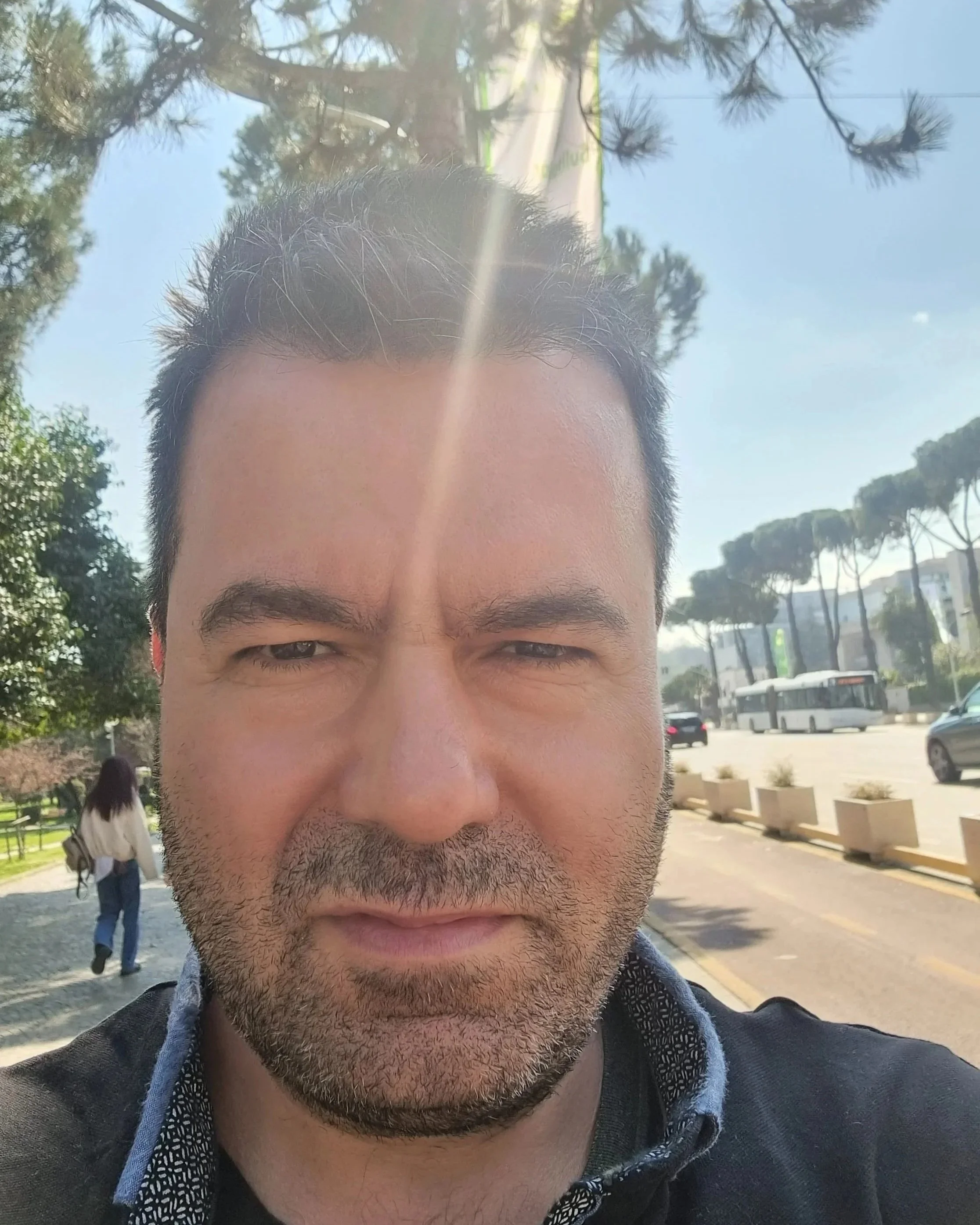 Un homme avec une barbe, prenant une selfie à l'extérieur par une journée ensoleillée, avec des arbres, une femme marchant, un bus, et une route en arrière-plan.