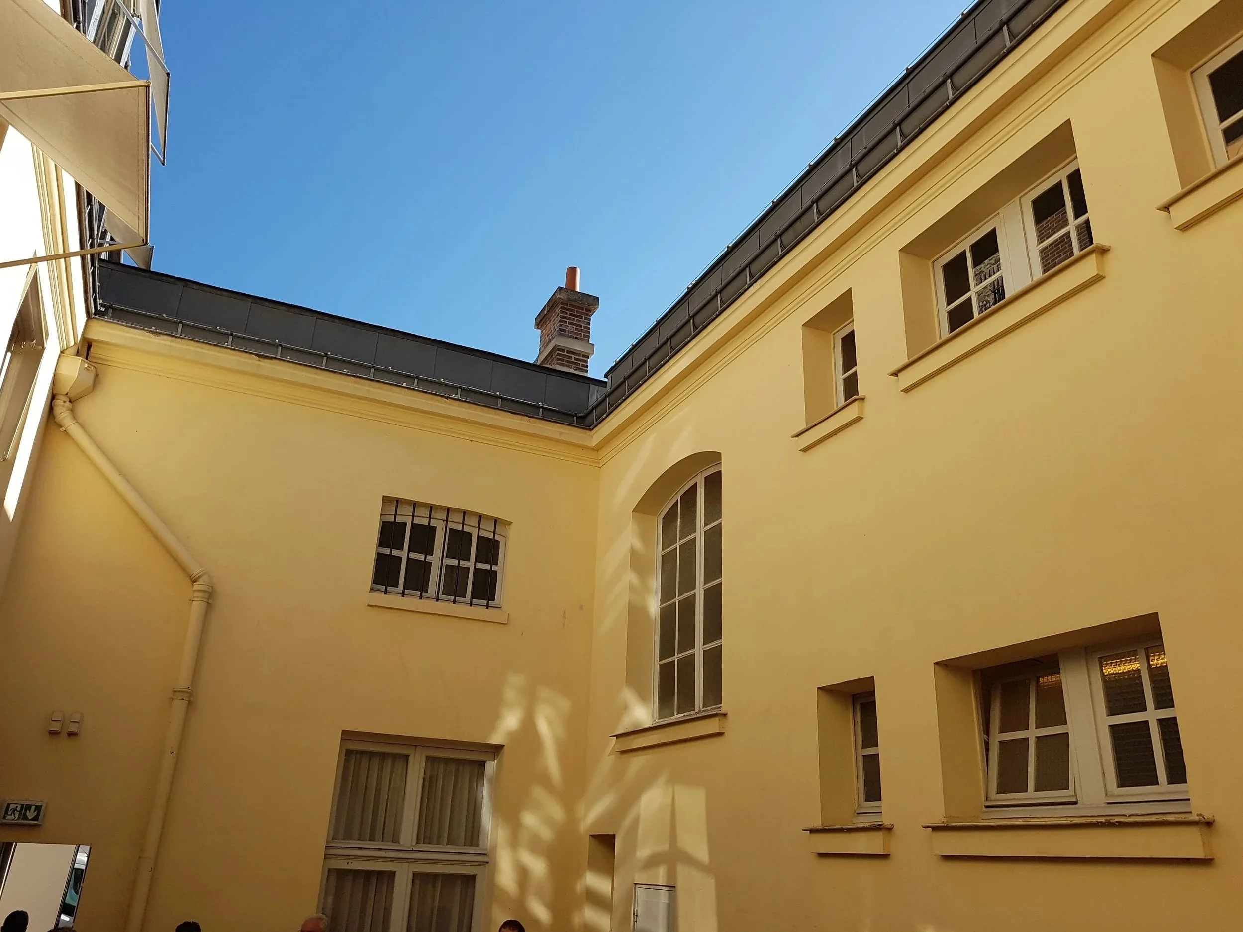 Vue du ciel d'une cour intérieure d'un bâtiment résidentiel de couleur jaune avec plusieurs fenêtres, un mur, une cheminée, un toit en ardoise et un ciel bleu clair
