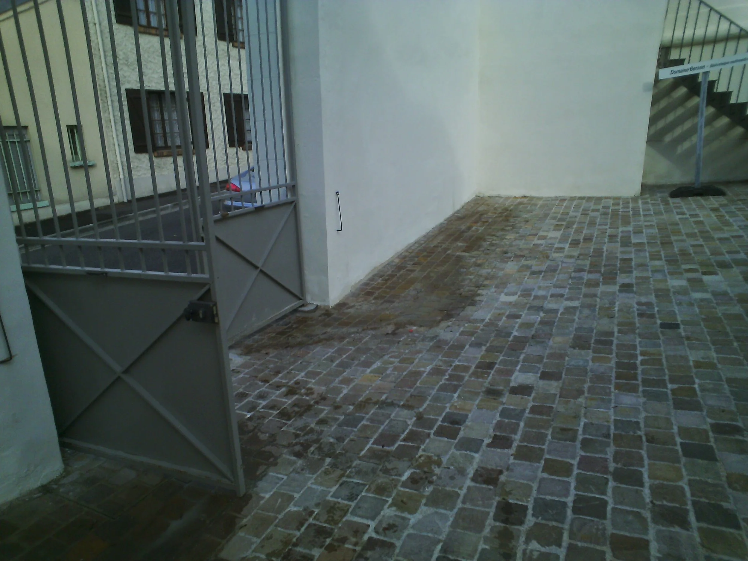 Un entrée de cour avec un portail métallique gris, un sol en pavés en terre cuite et un mur blanc. Il y a un escalier en métal en arrière-plan à droite.