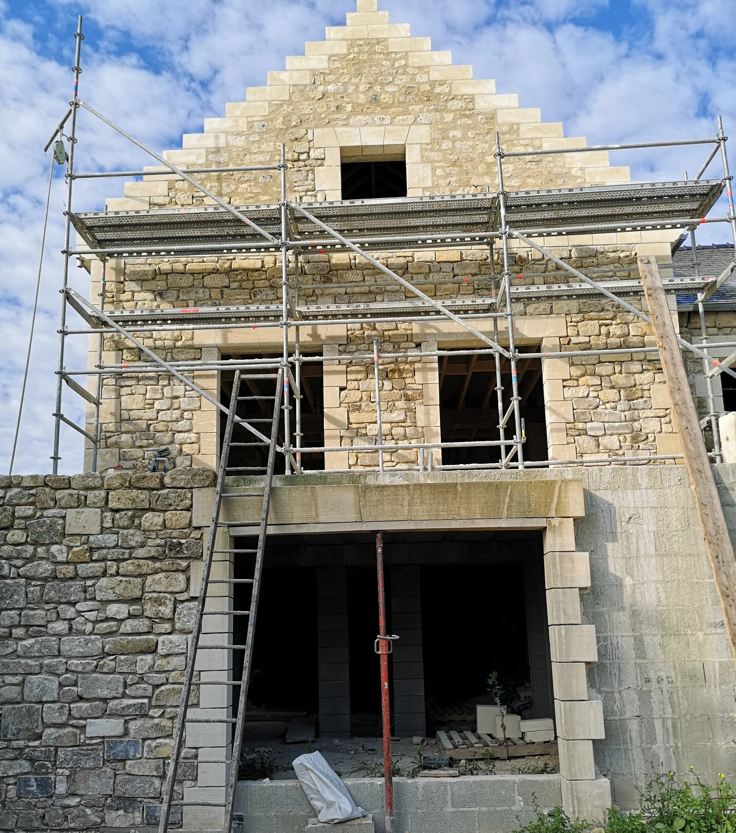 Construction d'une maison en pierre avec échafaudages autour, portes et fenêtres non encore installées, sous un ciel nuageux.